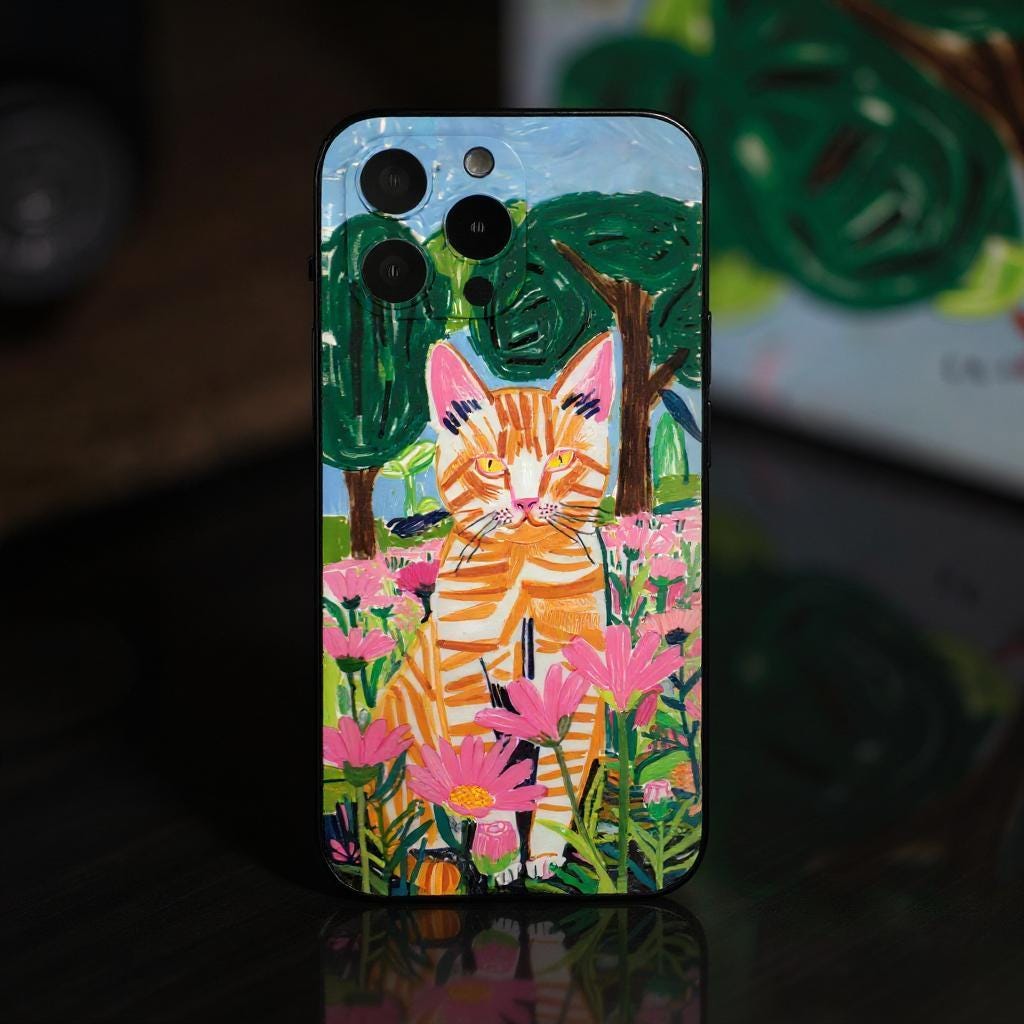 A custom pet portrait iPhone case featuring a vibrant illustration of an orange tabby cat in a field of pink flowers, set against a backdrop of green trees.