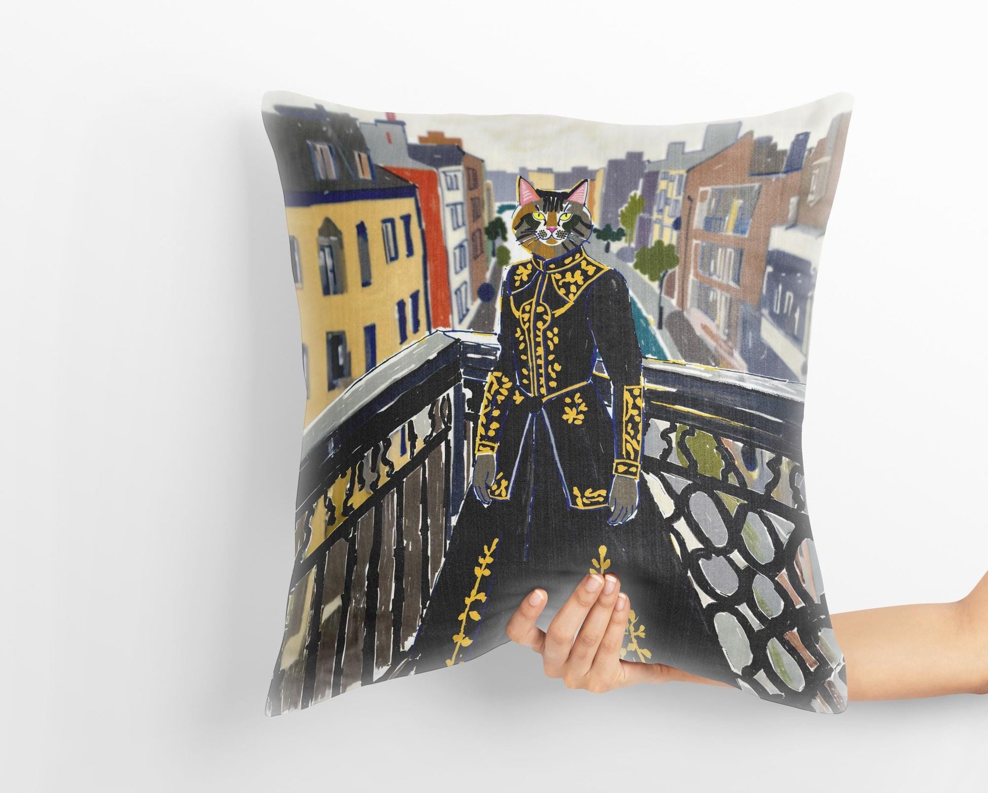 A hand holds a custom medieval pet portrait pillow featuring a tabby cat dressed in a regal black and gold gown, standing on a balcony overlooking a cityscape. The whimsical art adds a touch of humor and sophistication.