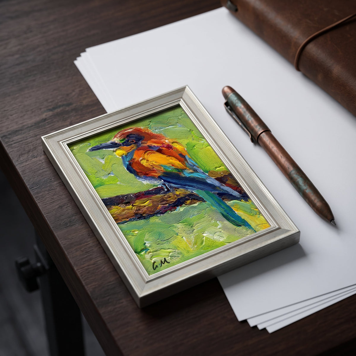 A vividly painted bird perched on a branch rests in a silver frame atop a dark wood desk, surrounded by paper and a copper pen.