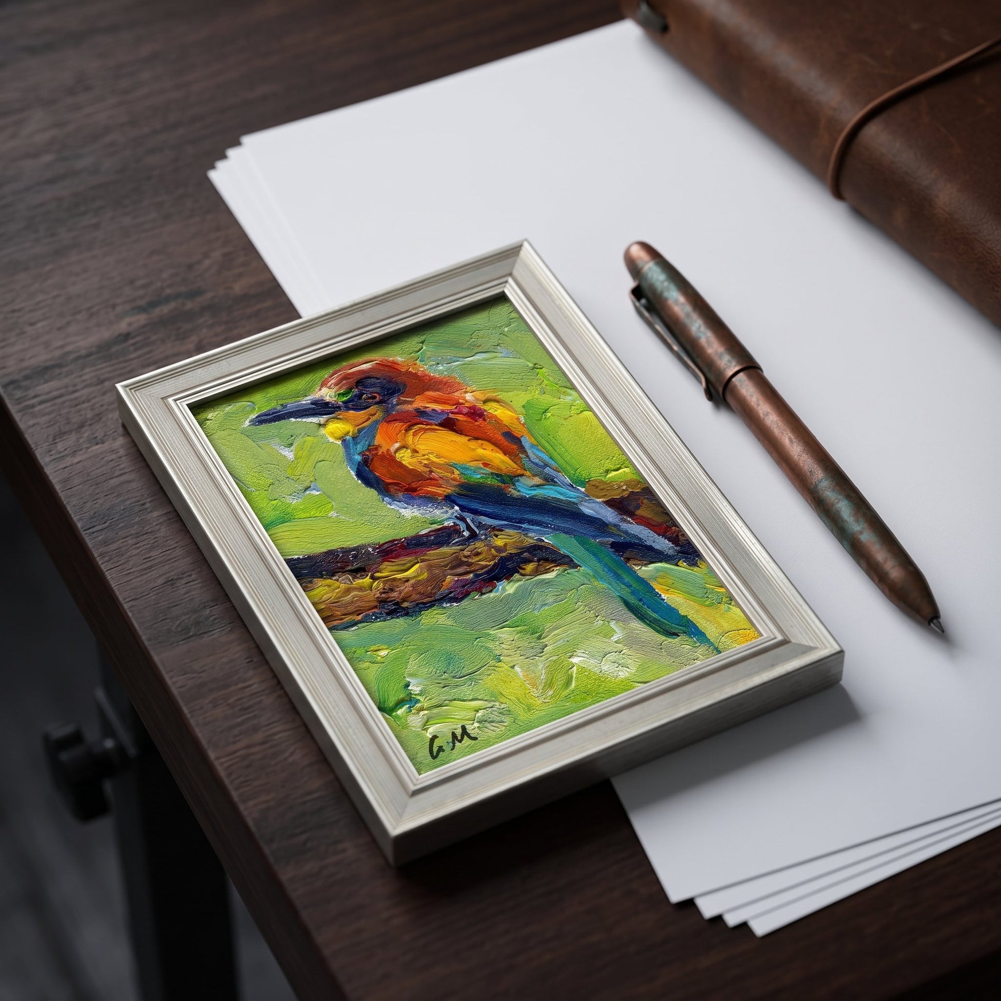 A vividly painted bird perched on a branch rests in a silver frame atop a dark wood desk, surrounded by paper and a copper pen.