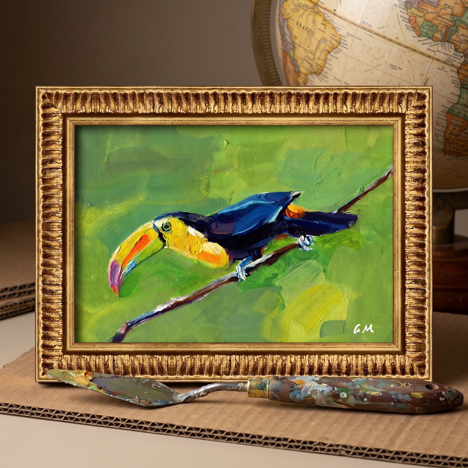A colorful toucan painting in a gold frame sits on a worktable beside a paint-stained palette knife and a globe, unframed canvas visible within.