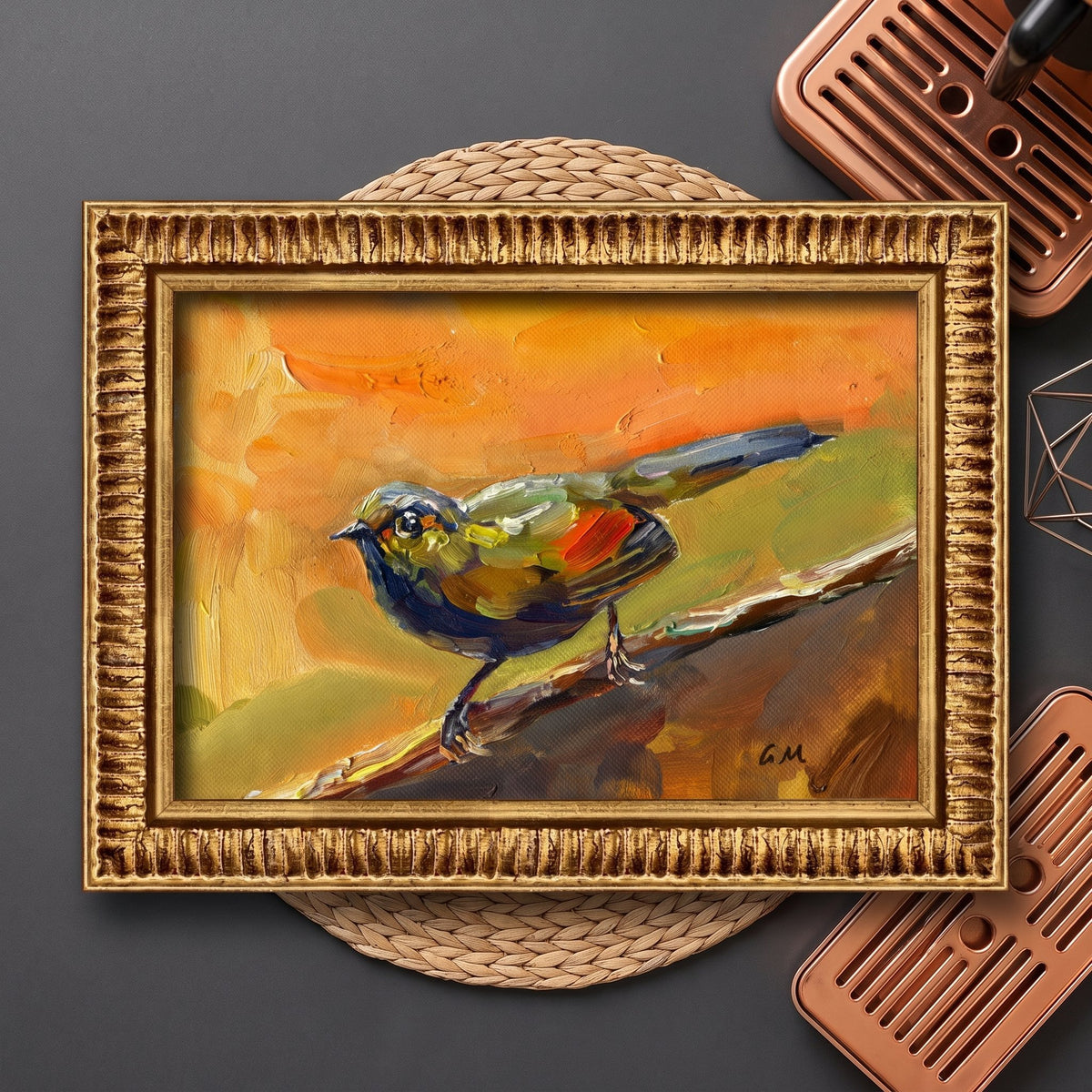 A vividly painted bird perched on a branch, rendered in bold orange, green, and blue tones, rests on a woven mat beside copper trays in a styled studio setting, framed in ornate gold.