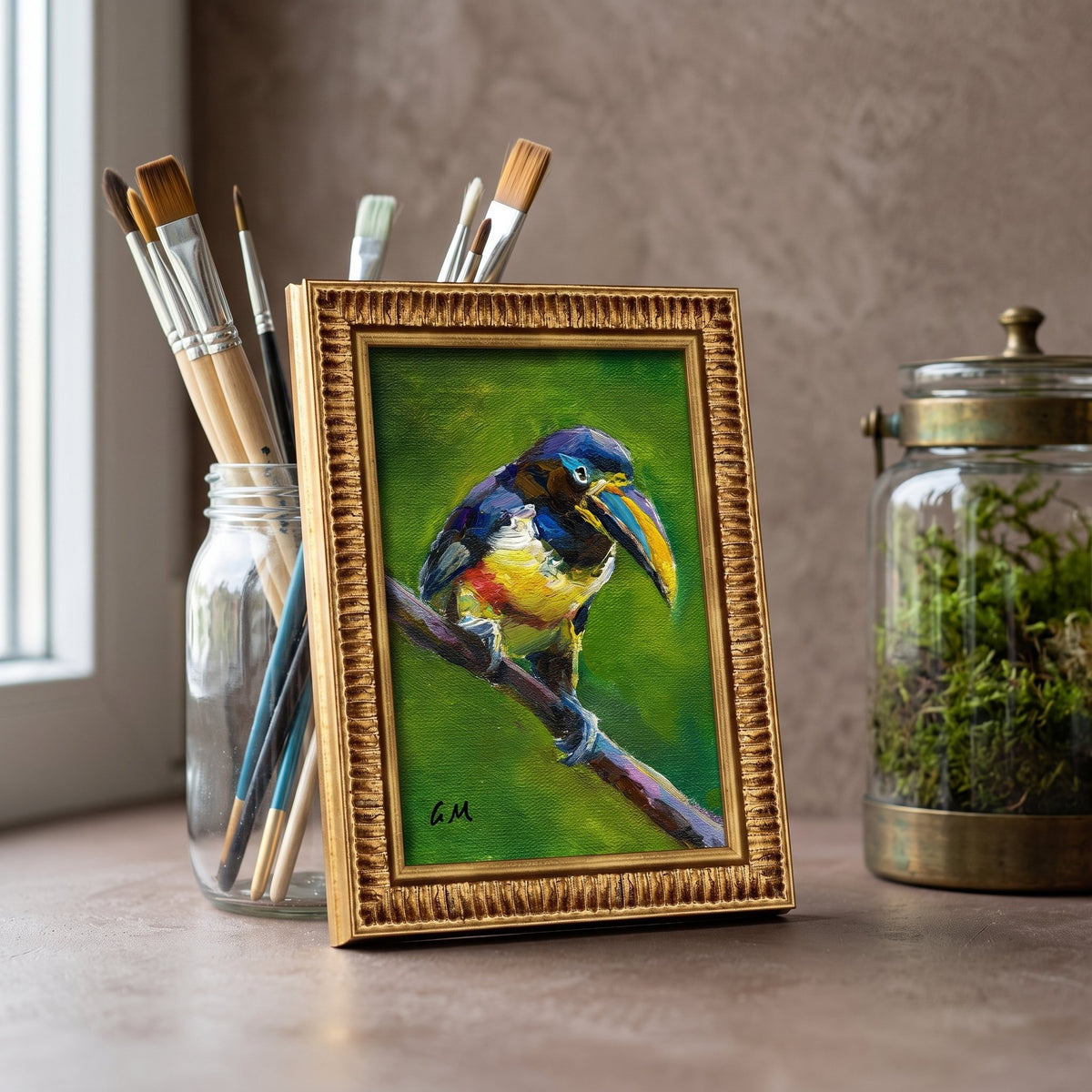 A colorful toucan painting in a gold frame rests on a studio counter beside paintbrushes and a moss terrarium, bathed in natural light.