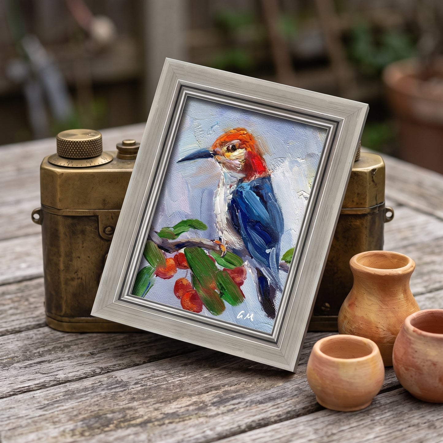 A vividly painted woodpecker perched on a branch with red berries, set in a silver frame on a rustic wooden table beside vintage camera gear and terracotta pots.