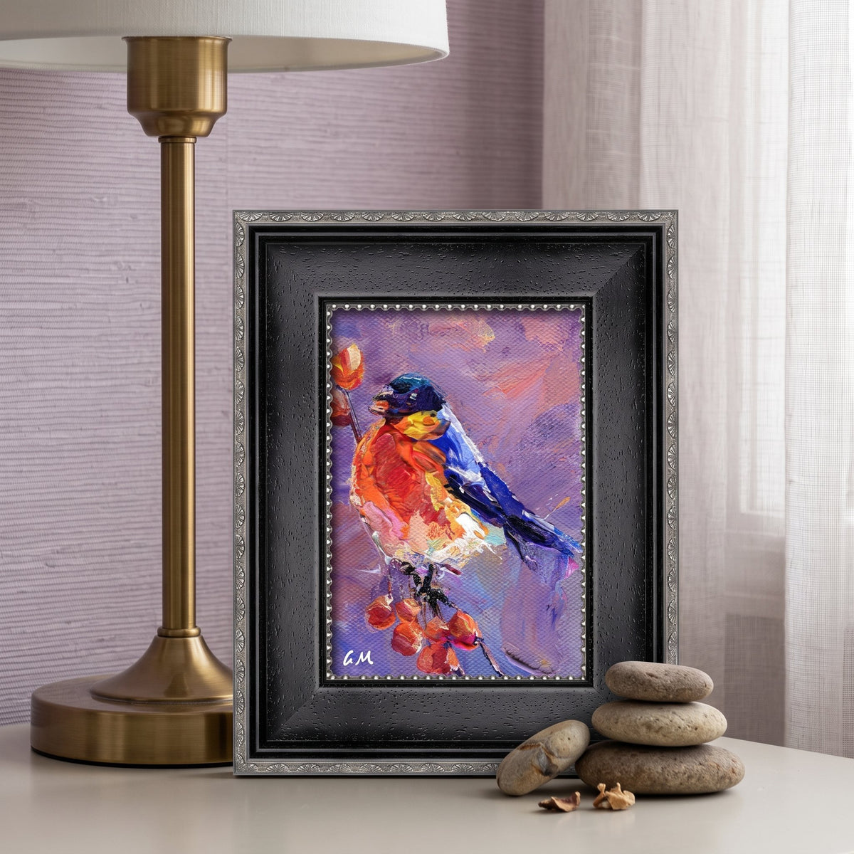 A vividly painted bird perched on berries in rich reds and blues, displayed in a black ornate frame on a bedside table beside a brass lamp and smooth stones.