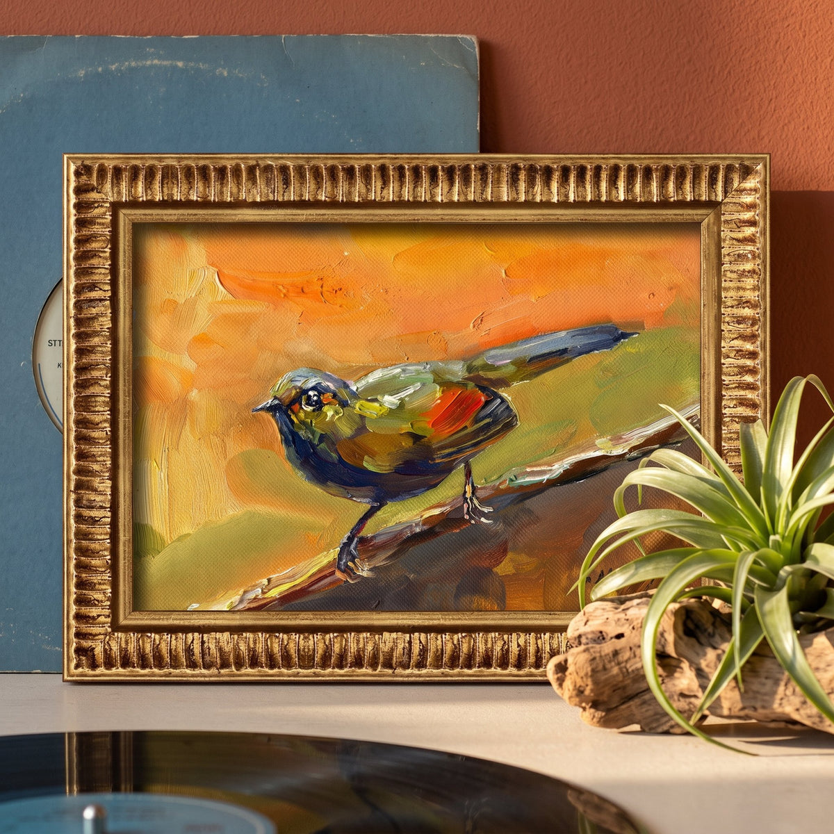 A vividly painted bird in orange, green, and blue perches on a branch in a golden-framed oil painting, displayed on a console beside a record player and air plant.