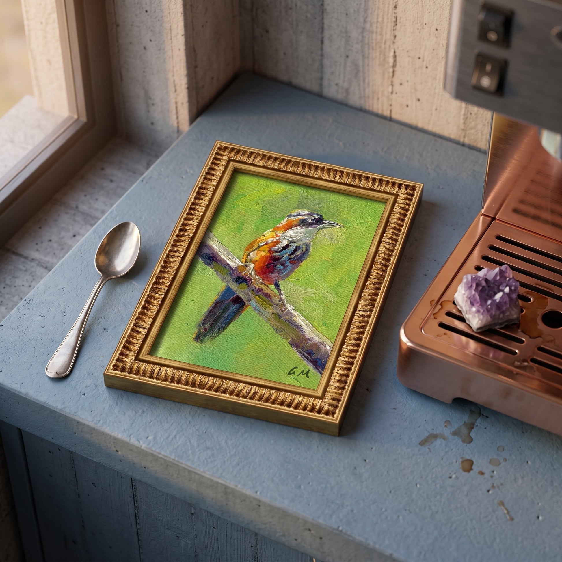 A vividly painted bird perched on a branch in warm greens and oranges, framed in gold, sits on a blue kitchen counter beside a coffee machine and spoon.