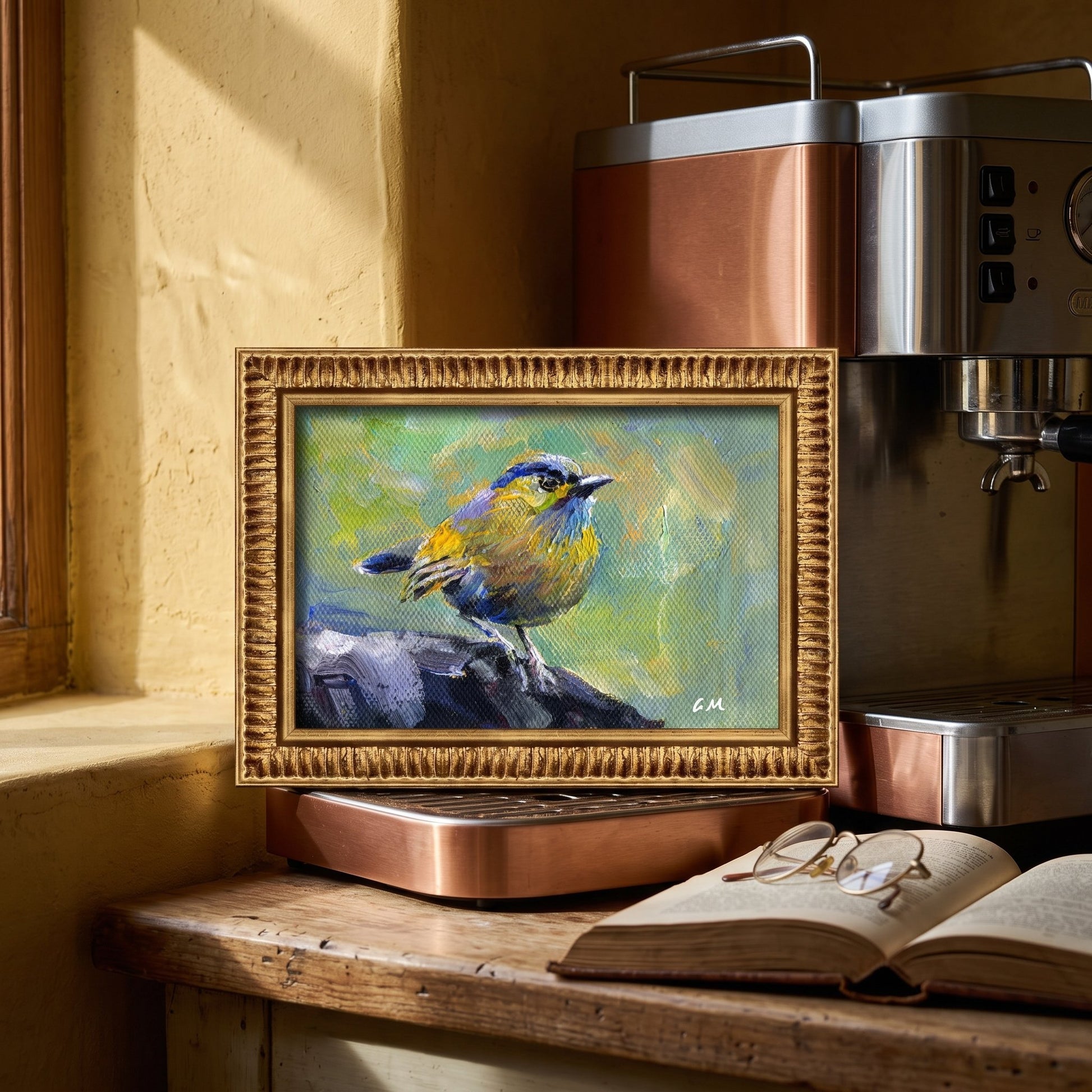 A vividly painted blue and yellow bird rests on a rock in a golden-framed canvas, nestled on a sunlit kitchen counter beside a coffee machine and open book.