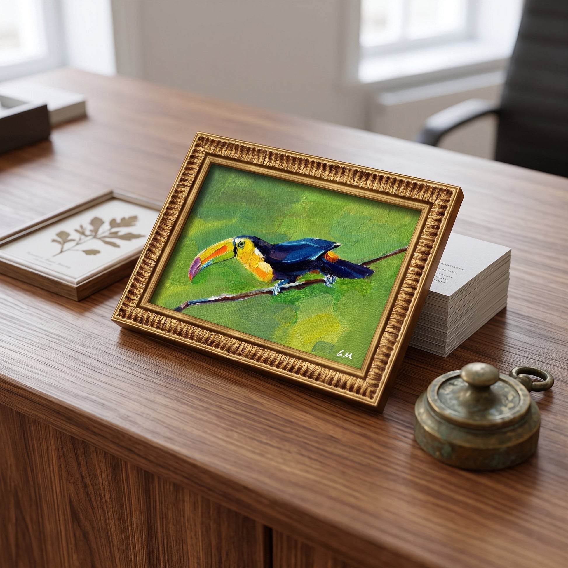 A colorful toucan painting in a gold frame sits on a wooden desk beside books and a vintage brass container, bathed in daylight.