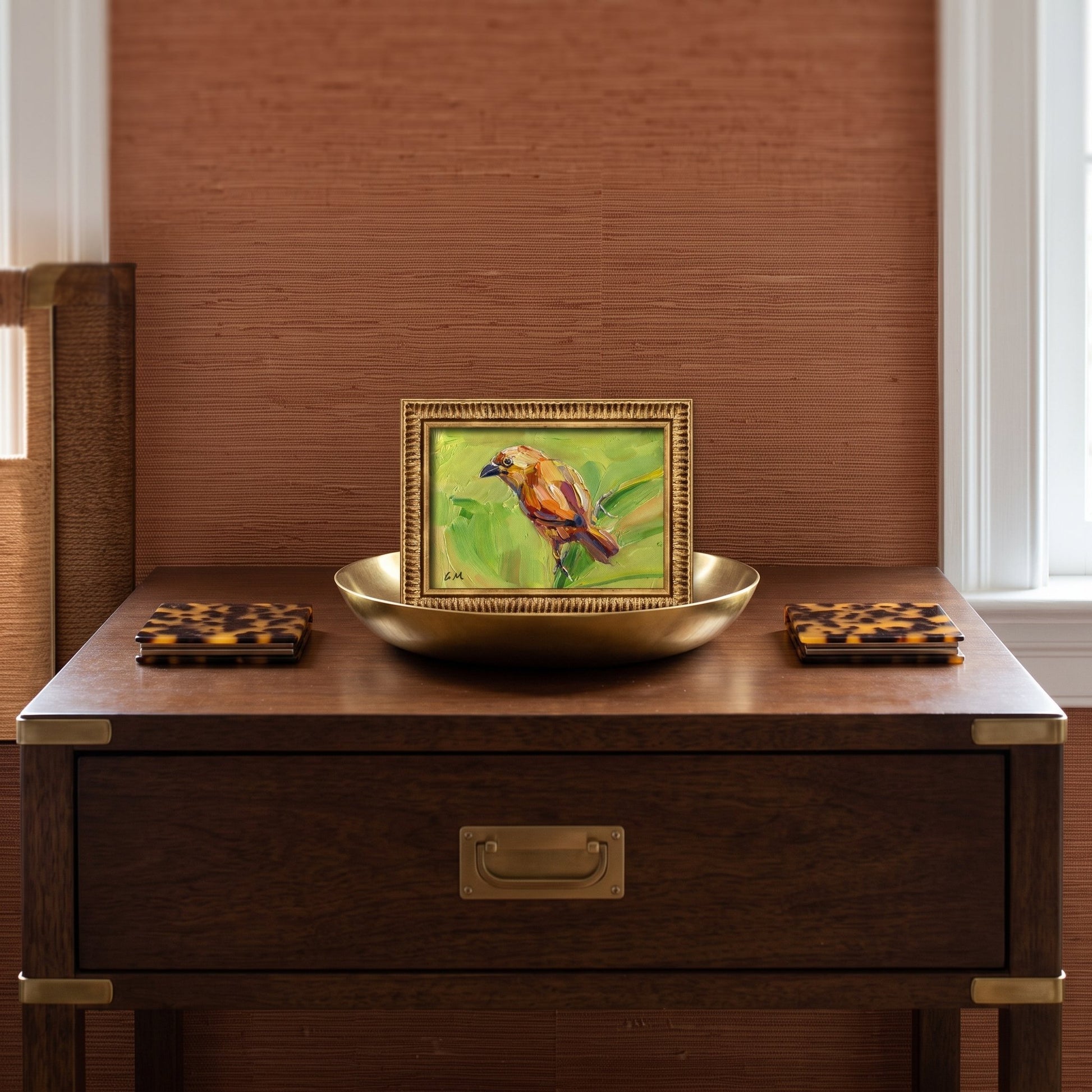 A golden-framed oil painting of a yellow bird on green leaves sits on a brass tray atop a dark wood console table in a sunlit study.