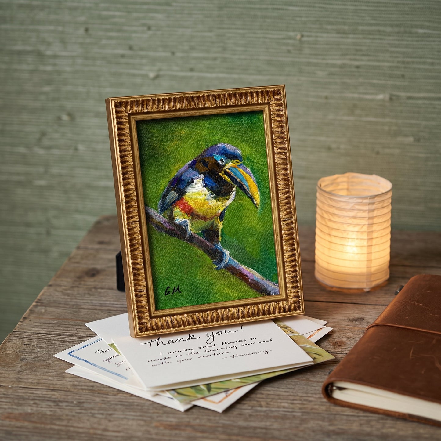 A colorful toucan painting in a gold frame rests on a wooden desk beside a glowing lantern and handwritten letters.