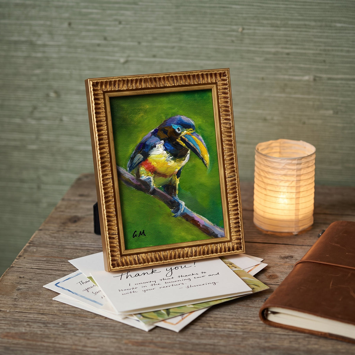 A colorful toucan painting in a gold frame rests on a wooden desk beside a glowing lantern and handwritten letters.