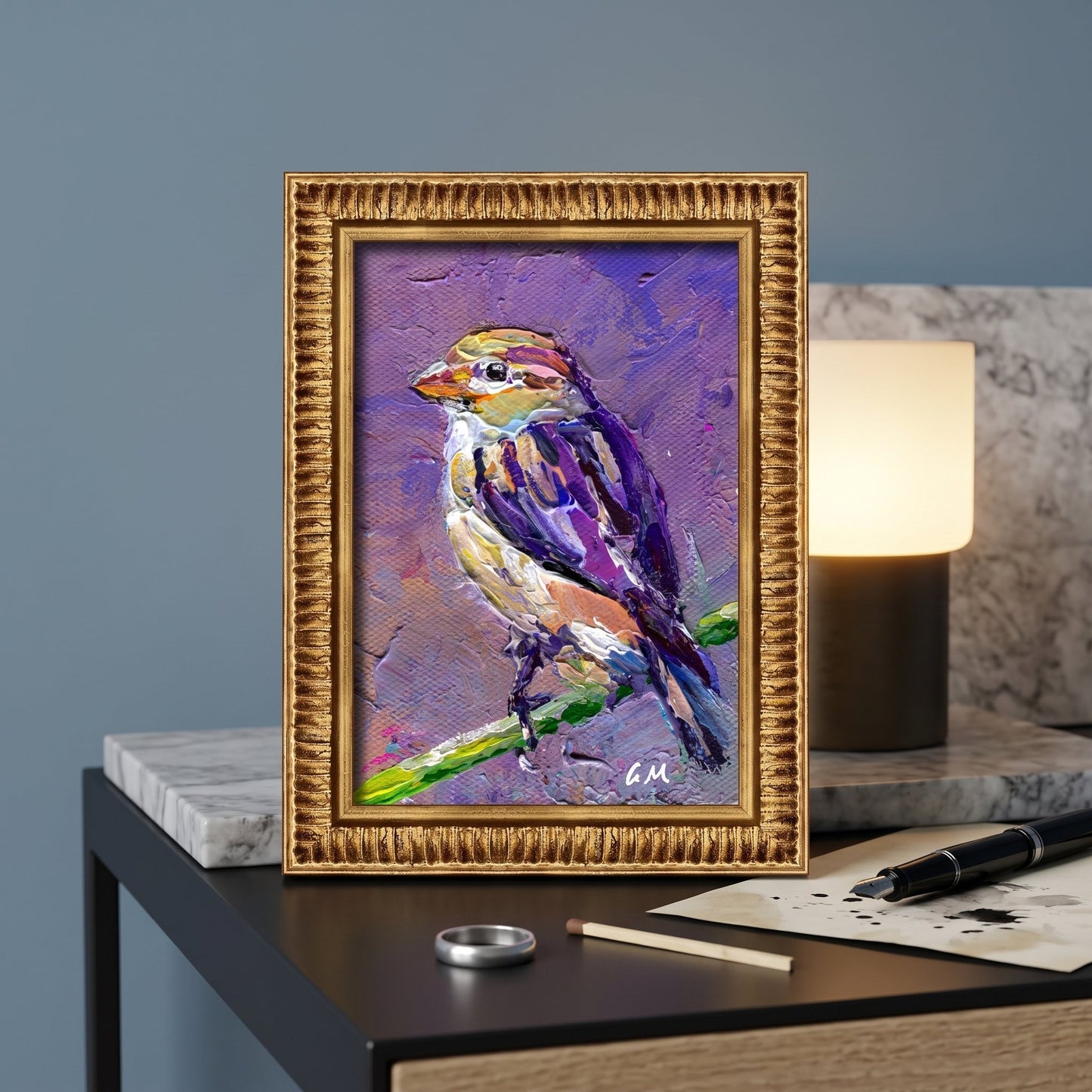 A vividly painted bird in purple and yellow hues perches on a green branch in a gold-framed canvas, displayed on a modern desk beside a glowing lamp.