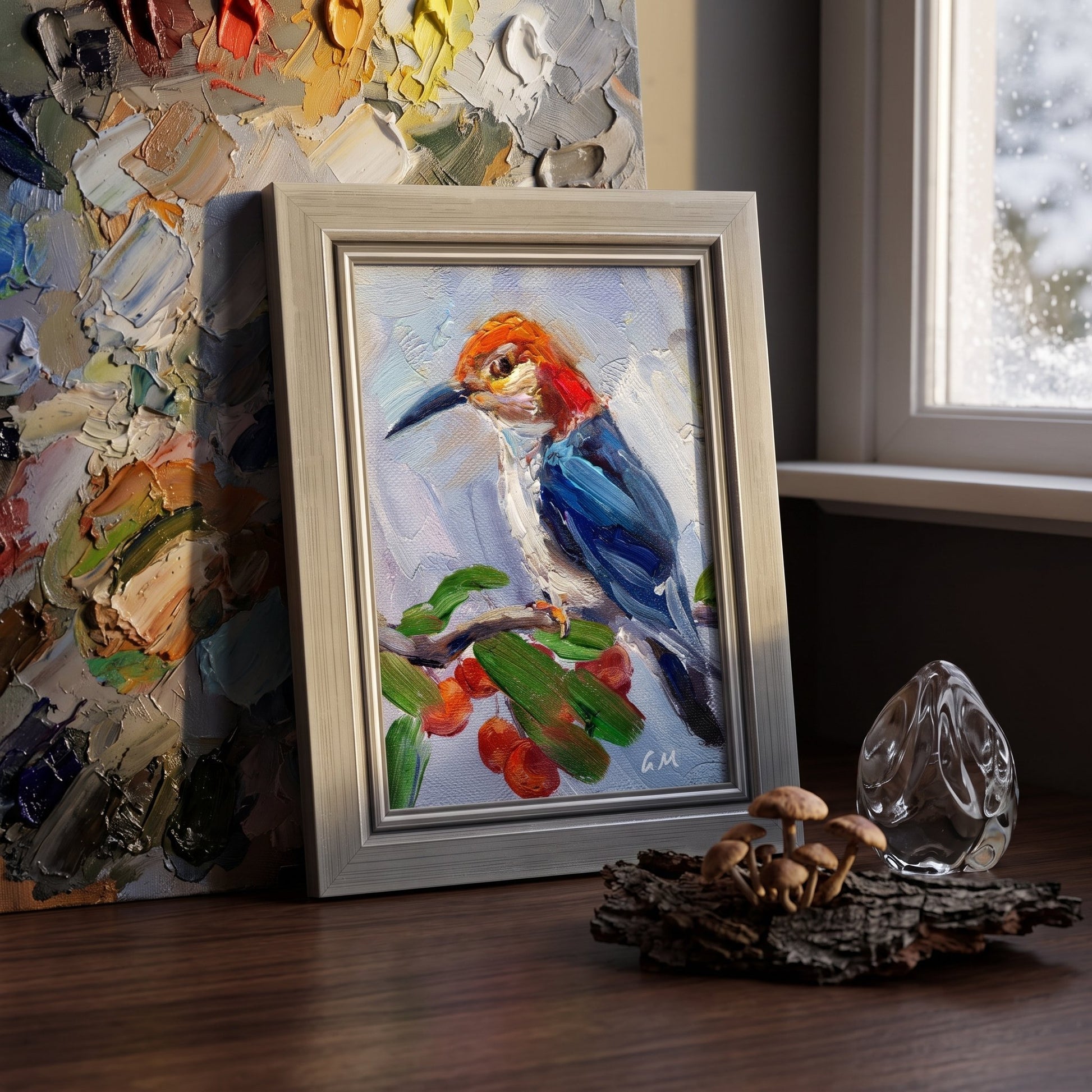 A vividly painted woodpecker with blue wings and red crest perches on a berry branch in a silver-framed canvas, set beside a painter's palette and natural decor on a wooden desk.