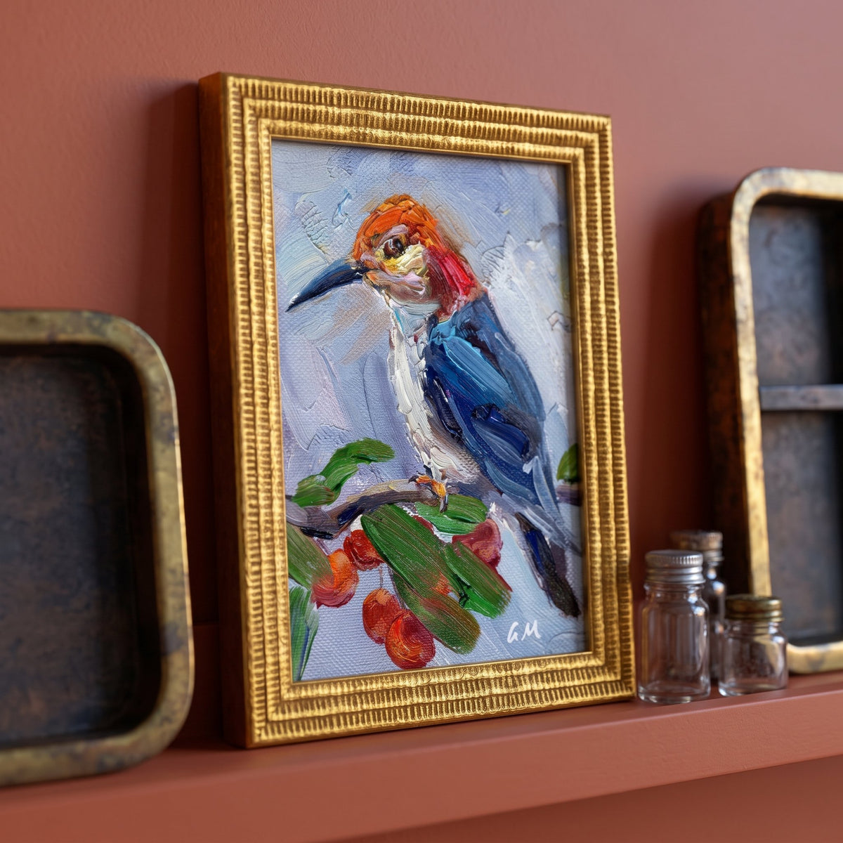 A colorful woodpecker painting in a gold frame rests on a terracotta shelf beside vintage jars and trays, unframed canvas visible beneath.