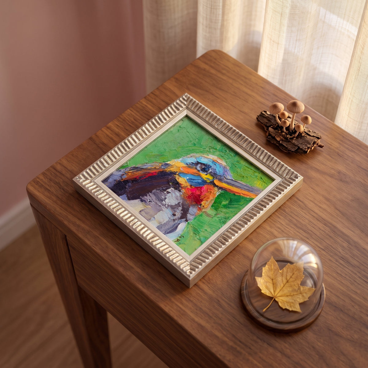 A colorful expressionist kingfisher painting in a silver frame sits on a wooden side table beside a glass dome with a maple leaf and a cluster of mushrooms.