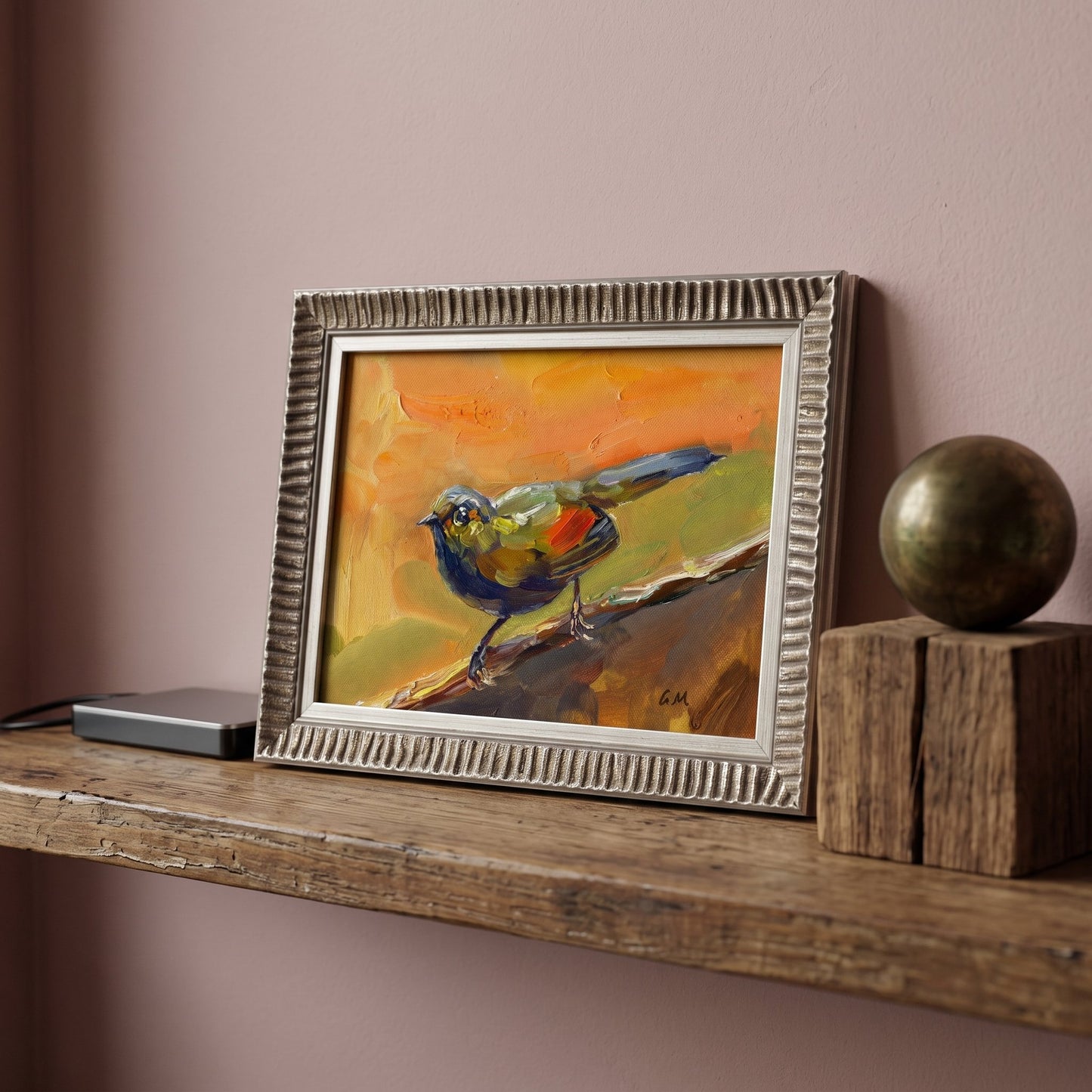 A colorful bird painting in a silver frame rests on a weathered wood shelf beside a brass orb, bathed in soft natural light.