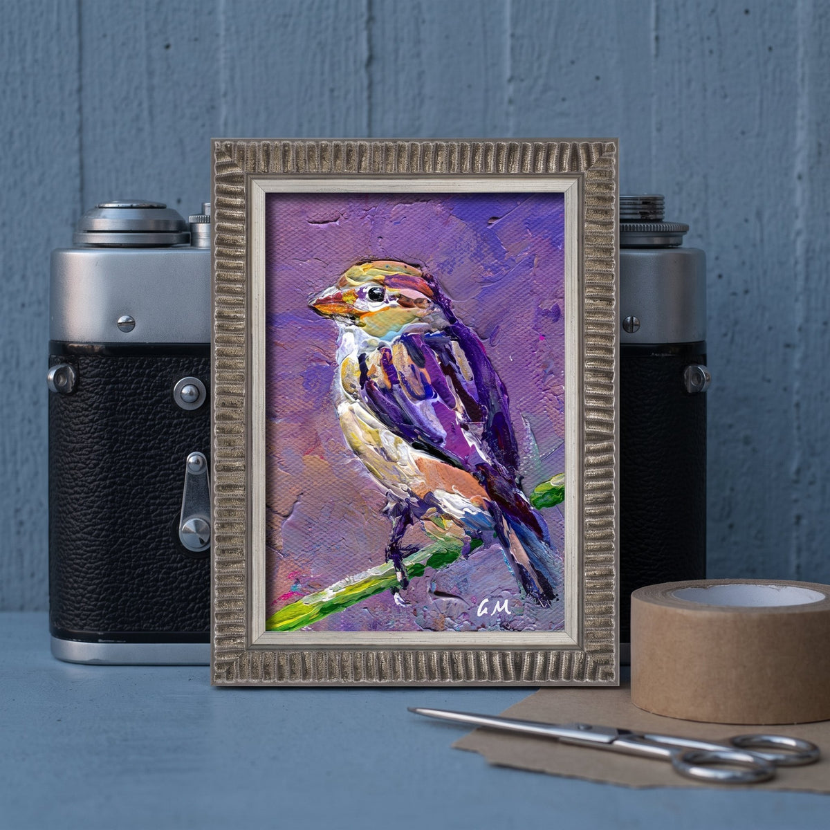 A vividly painted bird in purple and yellow hues perches on a green branch within a textured frame, set between vintage cameras on a blue workbench.