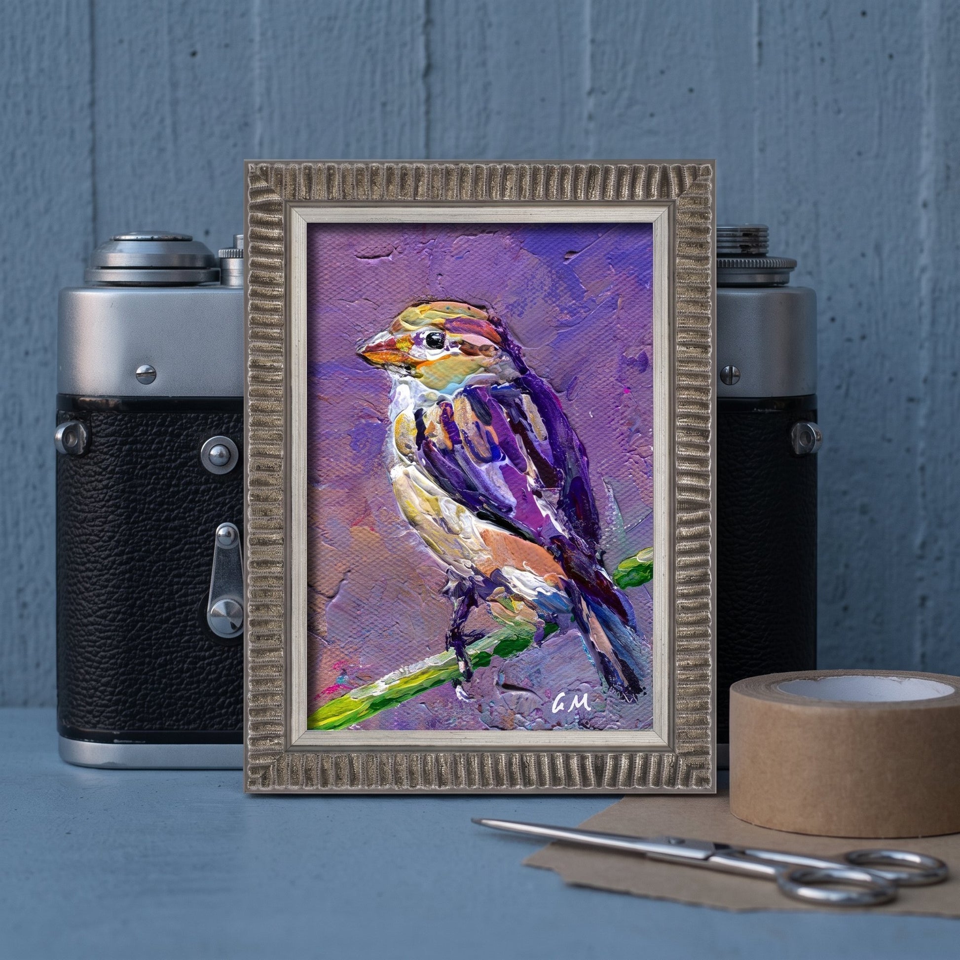 A vividly painted bird in purple and yellow hues perches on a green branch within a textured frame, set between vintage cameras on a blue workbench.
