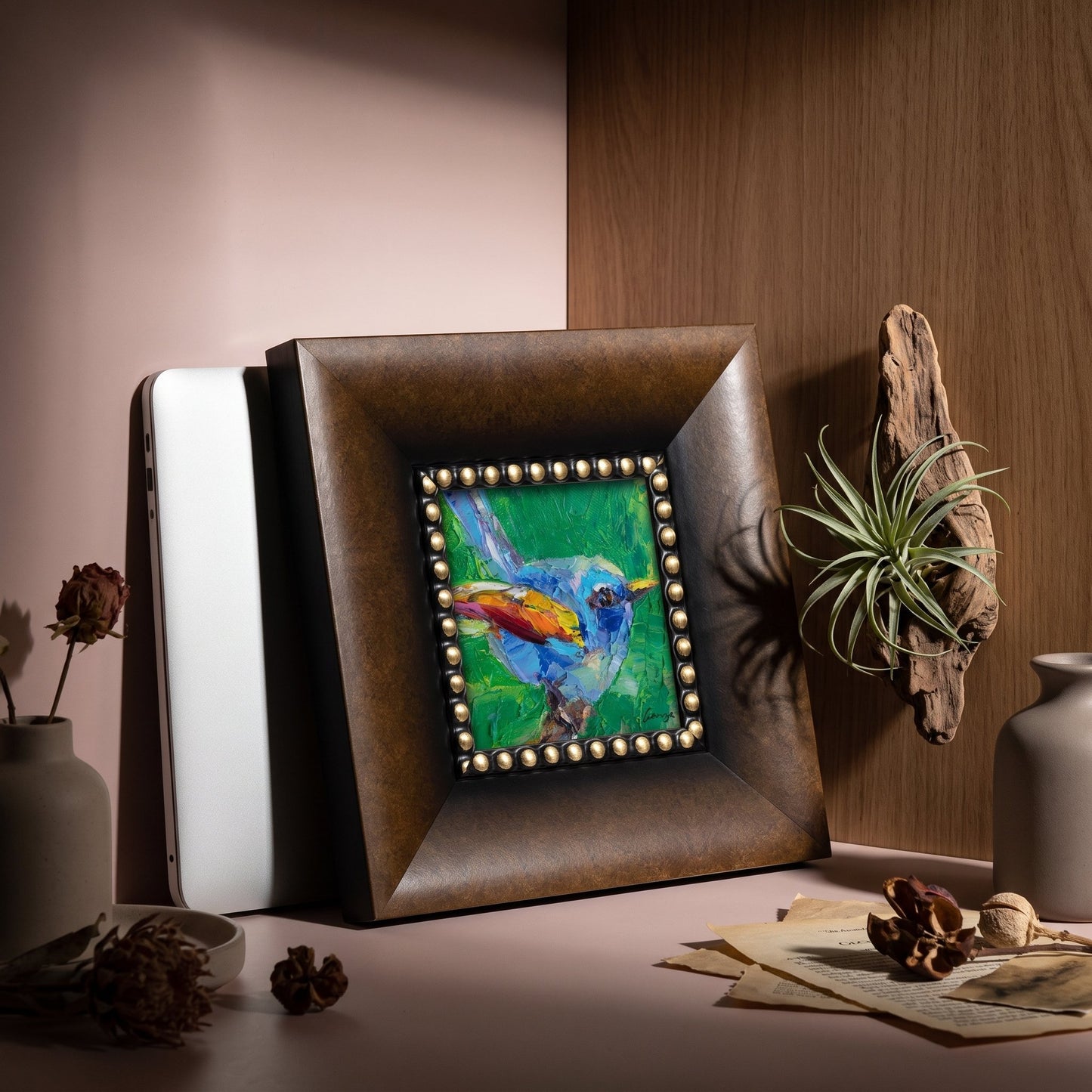 A vividly painted bird in flight on canvas rests in a leather-bound frame atop a softly lit desk, surrounded by natural textures and quiet study essentials.