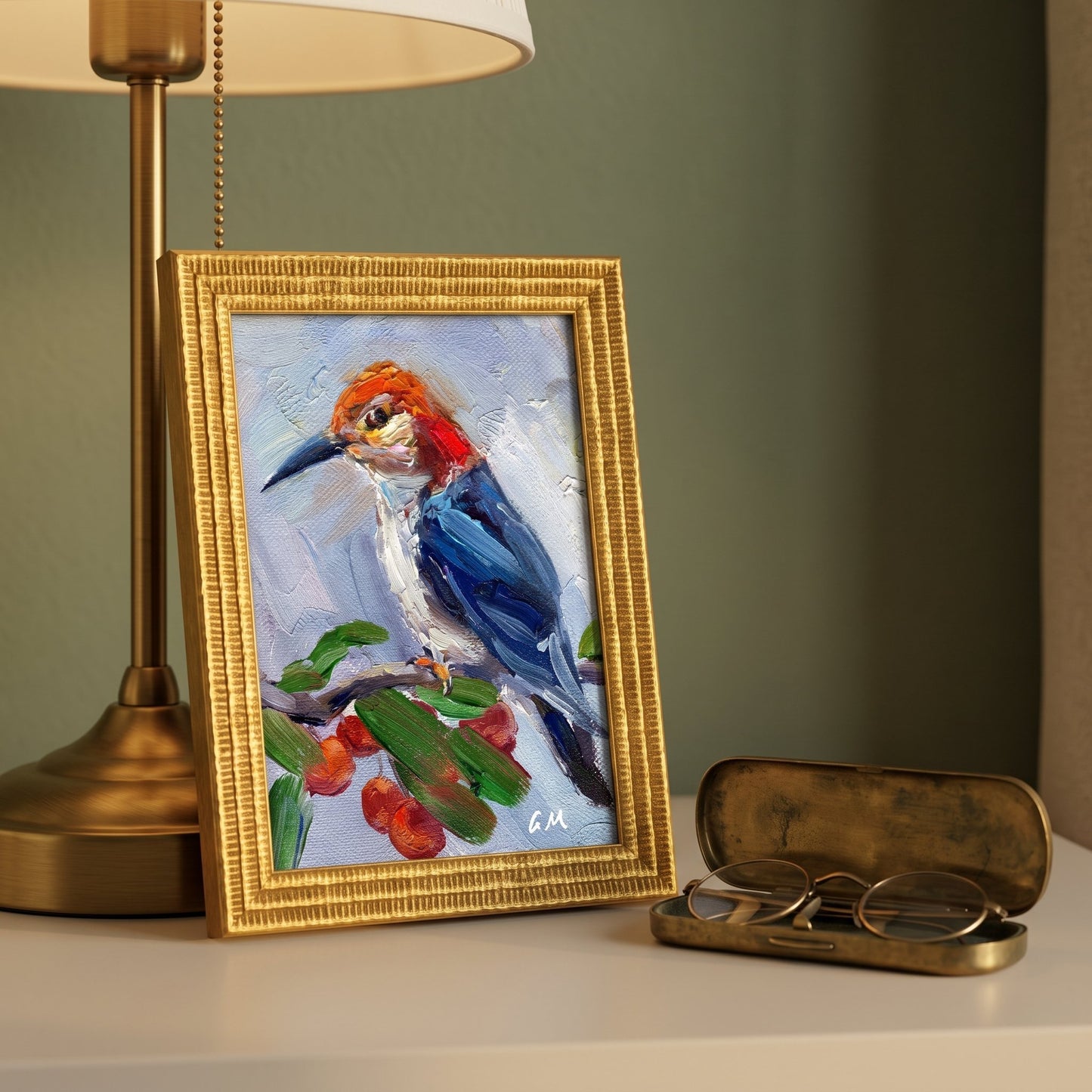 A colorful oil painting of a woodpecker perched on a berry-laden branch, displayed in a gold frame beside a brass lamp and vintage glasses case on a white desk.