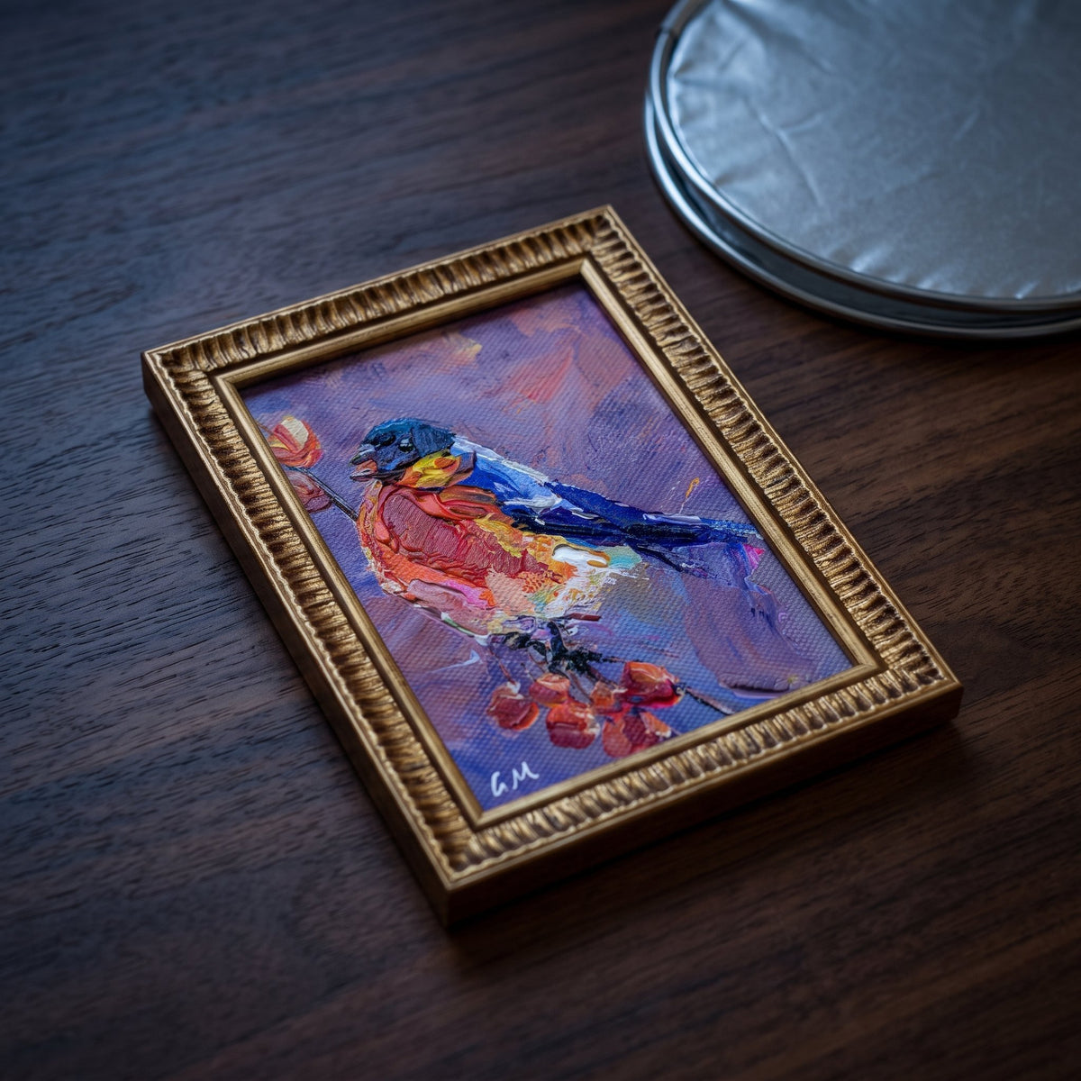 A vividly painted bird in red, blue, and purple hues rests on a dark wooden desk, framed in ornate gold, beside stacked silver coasters.