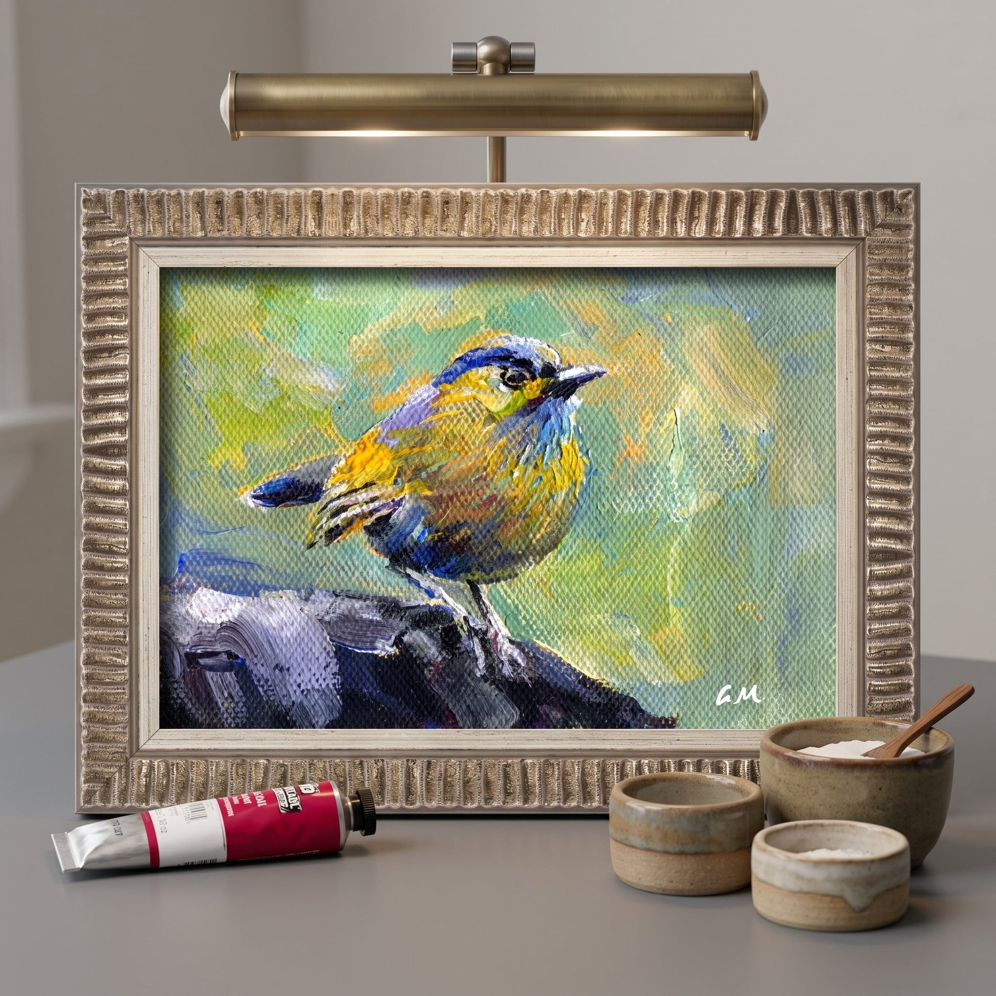 A vividly painted blue and yellow bird perches on a rock in an unframed canvas, illuminated by a brass desk lamp in a minimalist studio setting.