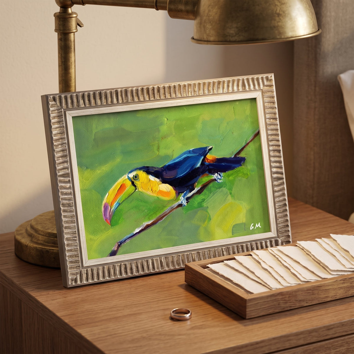 A colorful toucan painting perched on a branch, set against a green backdrop, sits on a wooden desk beside a brass lamp and a tray of notecards.