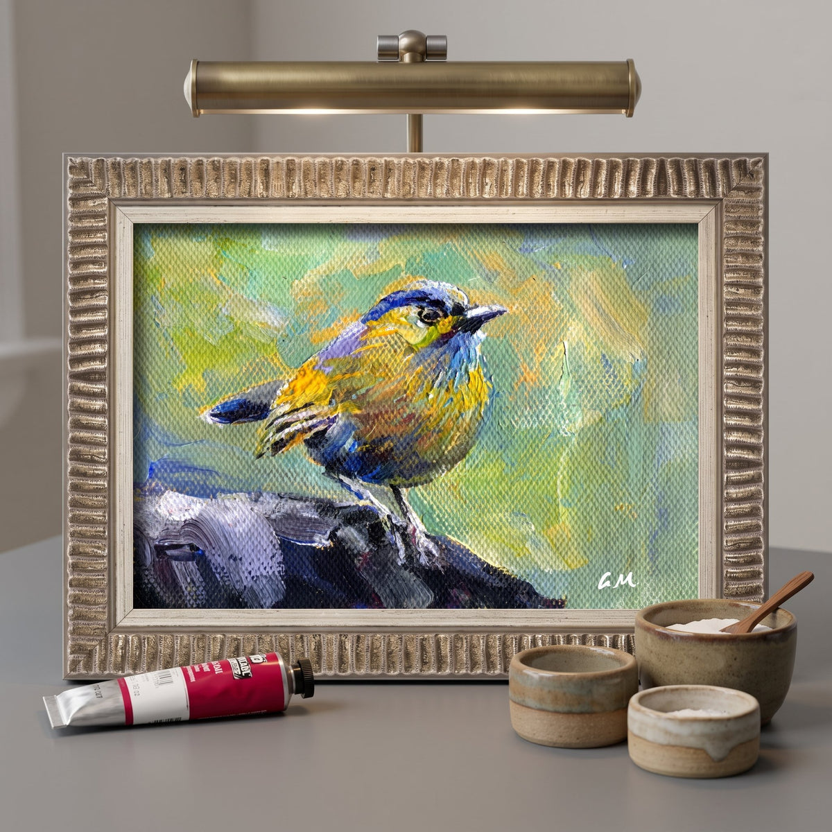 A vividly painted blue and yellow bird perches on a rock in an unframed canvas, illuminated by a brass desk lamp in a minimalist studio setting.