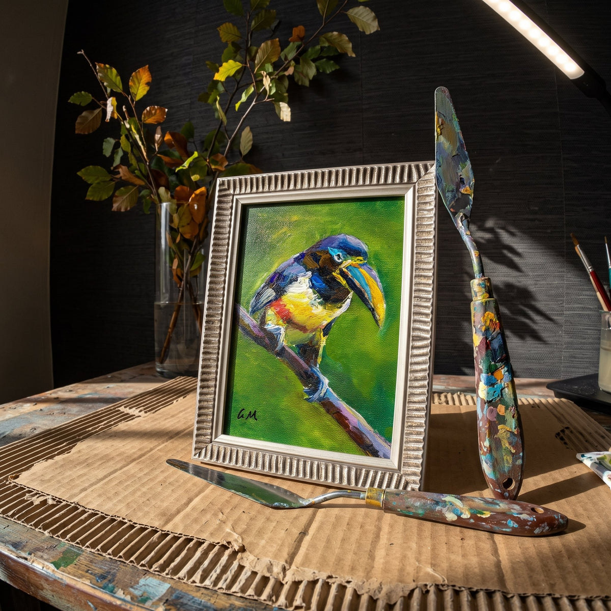 A vivid oil painting of a colorful toucan perched on a branch, framed in silver, rests on a paint-stained artist's worktable beside palette knives and dried leaves.