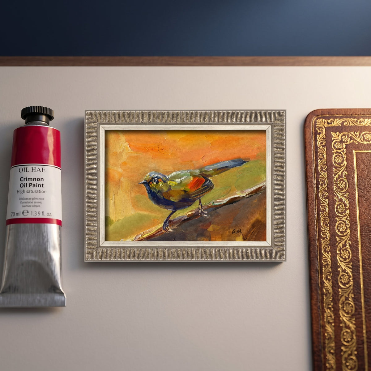 A vividly painted bird in orange, green, and blue rests on a branch in a silver-framed oil painting, displayed beside a tube of crimson oil paint and a vintage book in a curated artist's study.