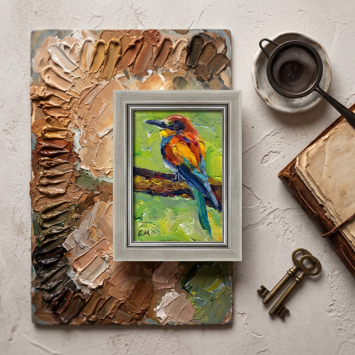 A vividly painted bird perched on a branch, rendered in bold oil strokes, rests within a silver frame atop a textured artist's palette in a sunlit studio setting.