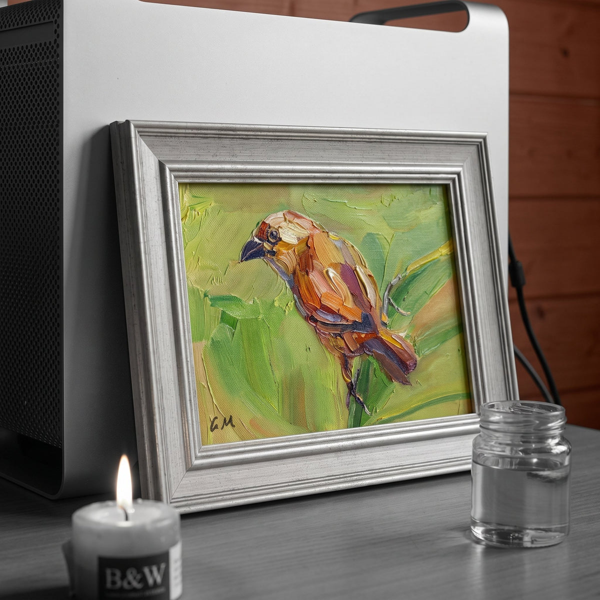 A colorful oil painting of a bird on canvas leans against a speaker on a desk, softly lit by a candle beside it.