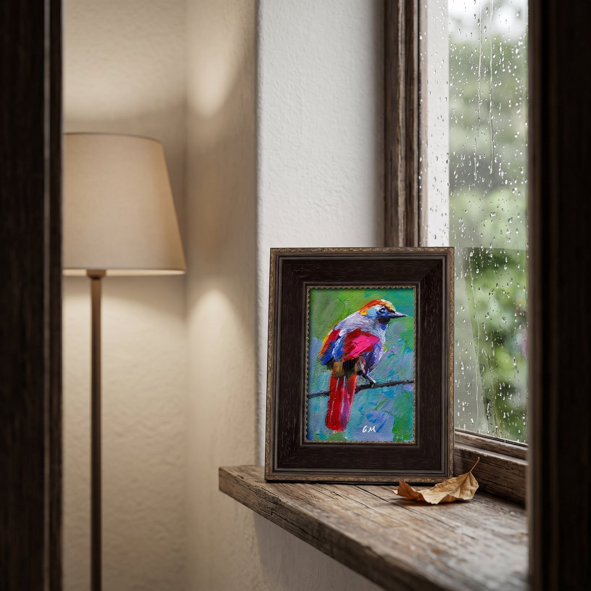 A vividly colored bird painting rests on a rain-speckled windowsill, framed in dark wood, with soft lamplight glowing beside it.