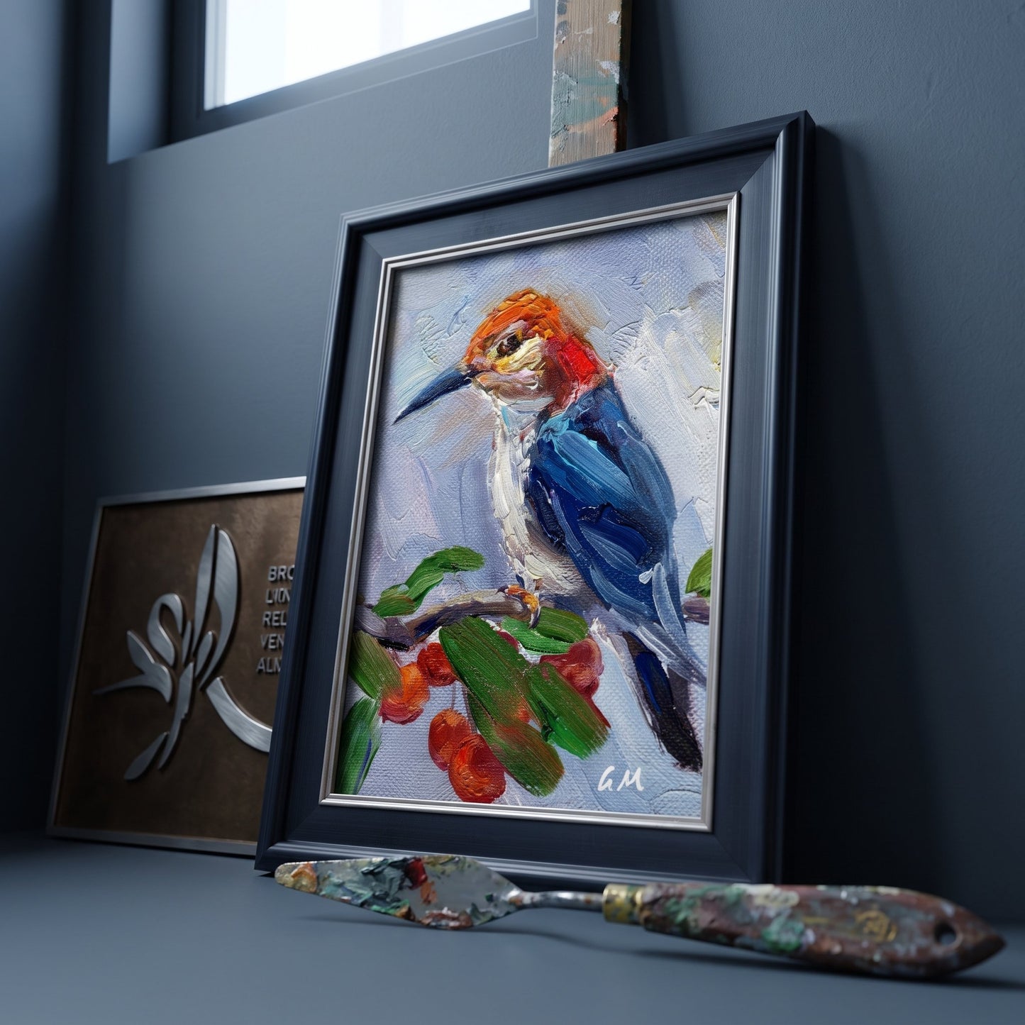 A vividly painted kingfisher perches on a berry-laden branch in a black-framed canvas, set against a dark studio wall beside an artist's palette knife.