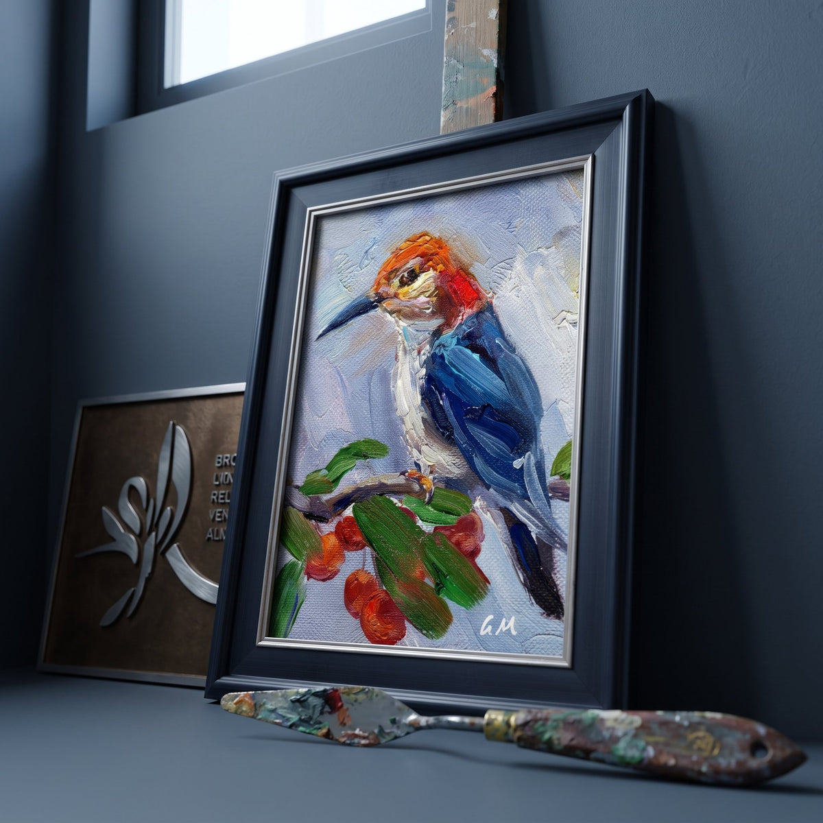 A vividly painted kingfisher perches on a berry-laden branch in a black-framed canvas, set against a dark studio wall beside an artist's palette knife.