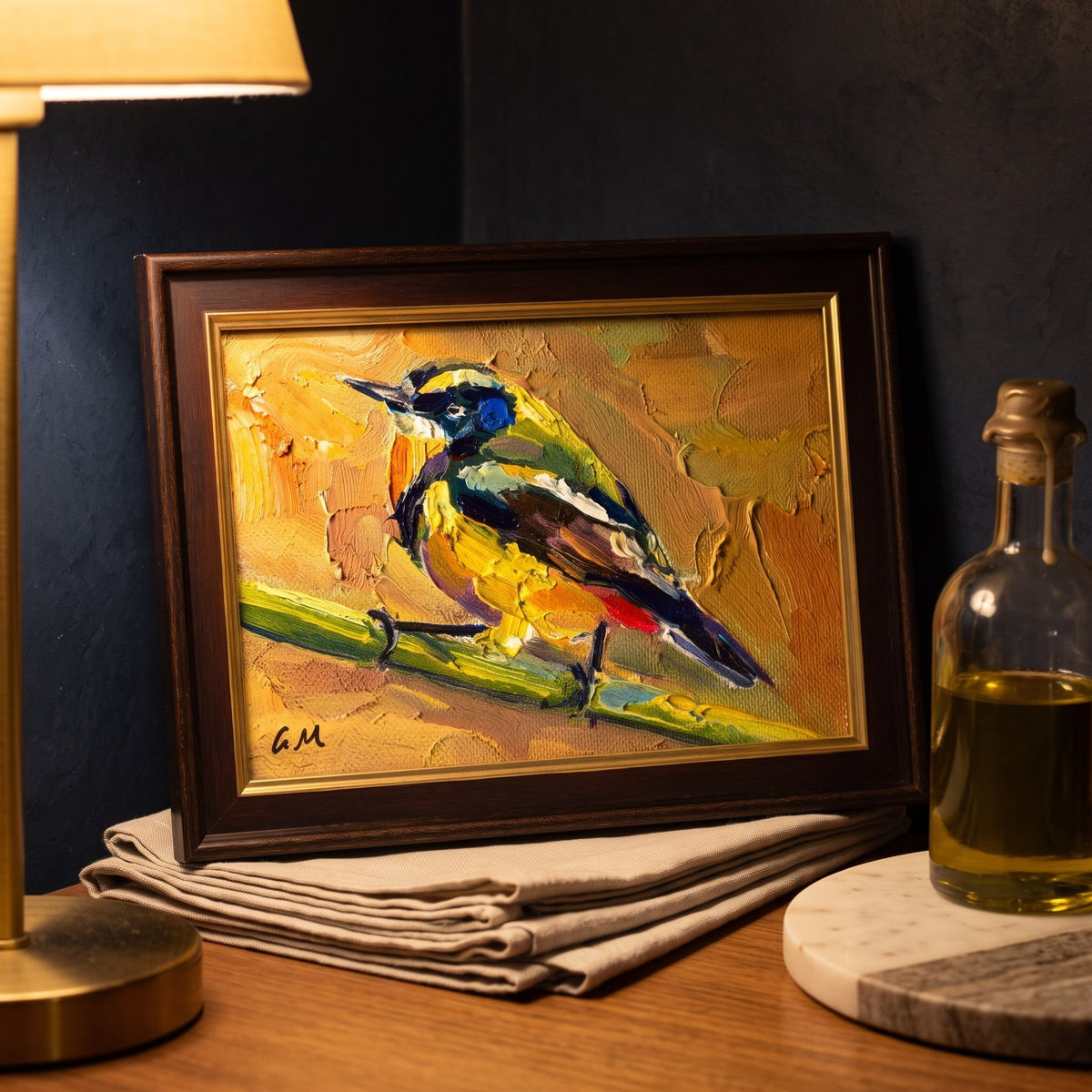 A vividly painted bird with yellow, blue, and brown plumage rests on a branch in a warmly lit study, displayed in a dark wooden frame atop a folded linen cloth.