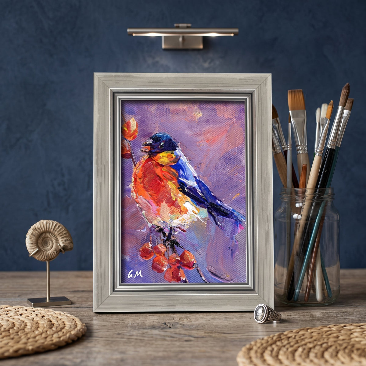 A vividly painted bird perched on berries in bold reds and blues rests on a wooden desk in an artist’s studio, unframed and glowing under warm light.
