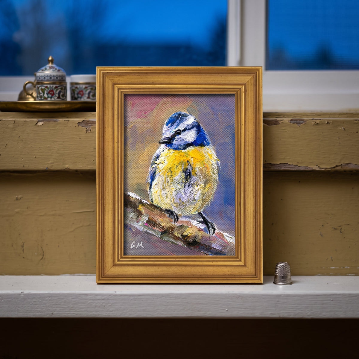 A vivid blue and yellow bird painting rests on a sunlit windowsill in a rustic farmhouse study, framed in gold.