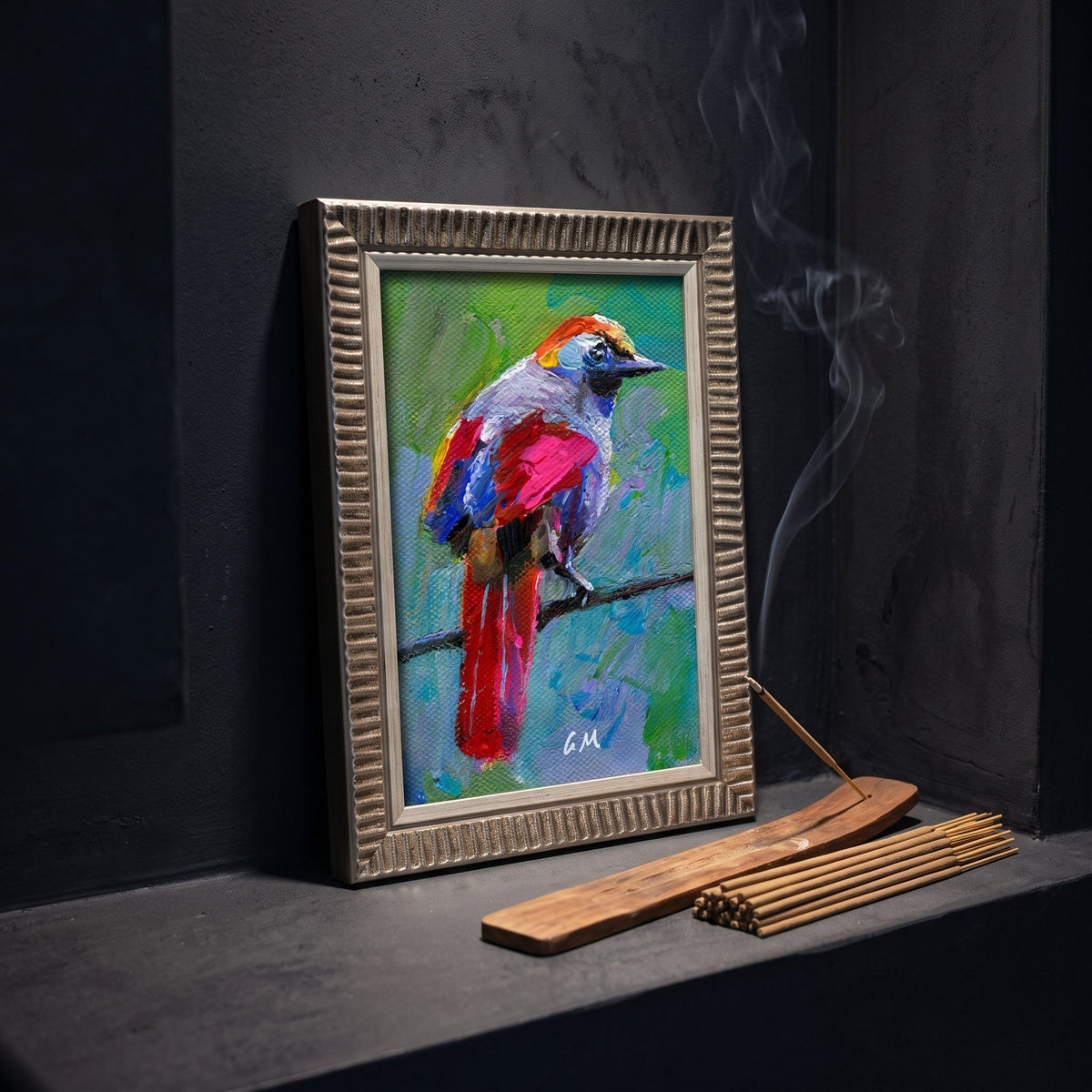 A vividly painted bird perched on a branch, bursting with red, blue, and green hues, leans against a dark wall beside a smoldering incense stick in a wooden holder—framed in textured silver.