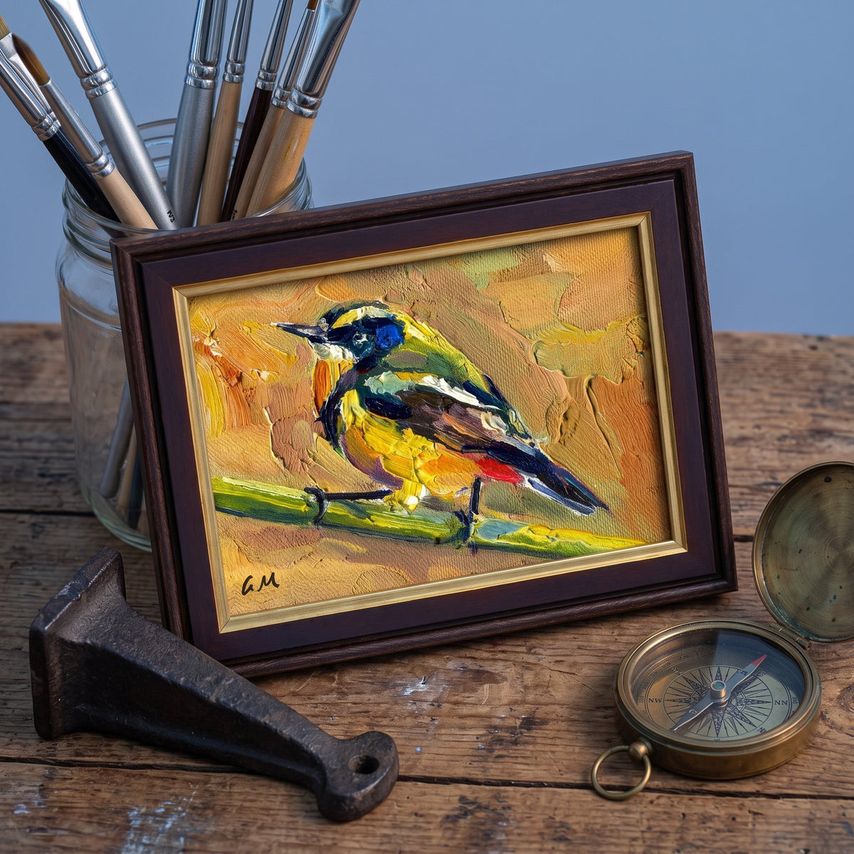 A vividly painted bird in yellow, blue, and brown rests on a branch in a framed canvas, set on a rustic wooden desk beside brushes and a compass.