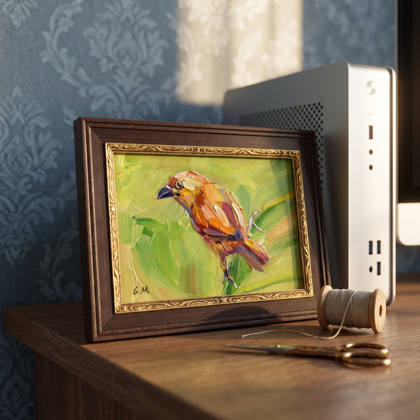 A vividly painted bird in warm earth tones rests on a wooden desk beside a spool of thread and scissors, framed in dark wood with gold detailing.