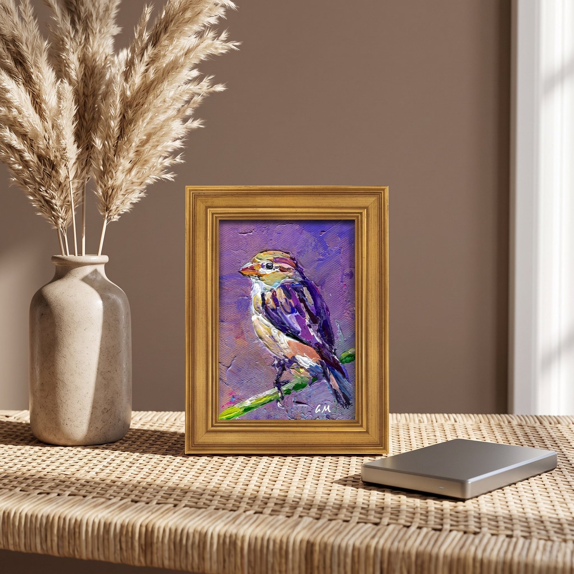 A vividly painted bird perched on a branch in purple and yellow hues sits in a gold frame on a woven table beside dried grasses, bathed in natural light.