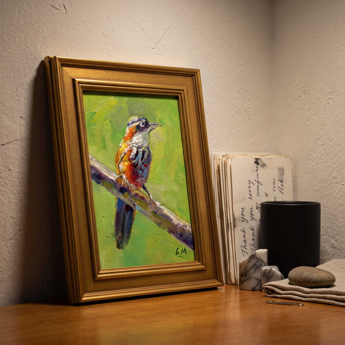 A vividly painted bird perched on a branch in warm hues of orange, green, and brown, displayed in a gold frame on a wooden desk beside handwritten notes and a black mug.
