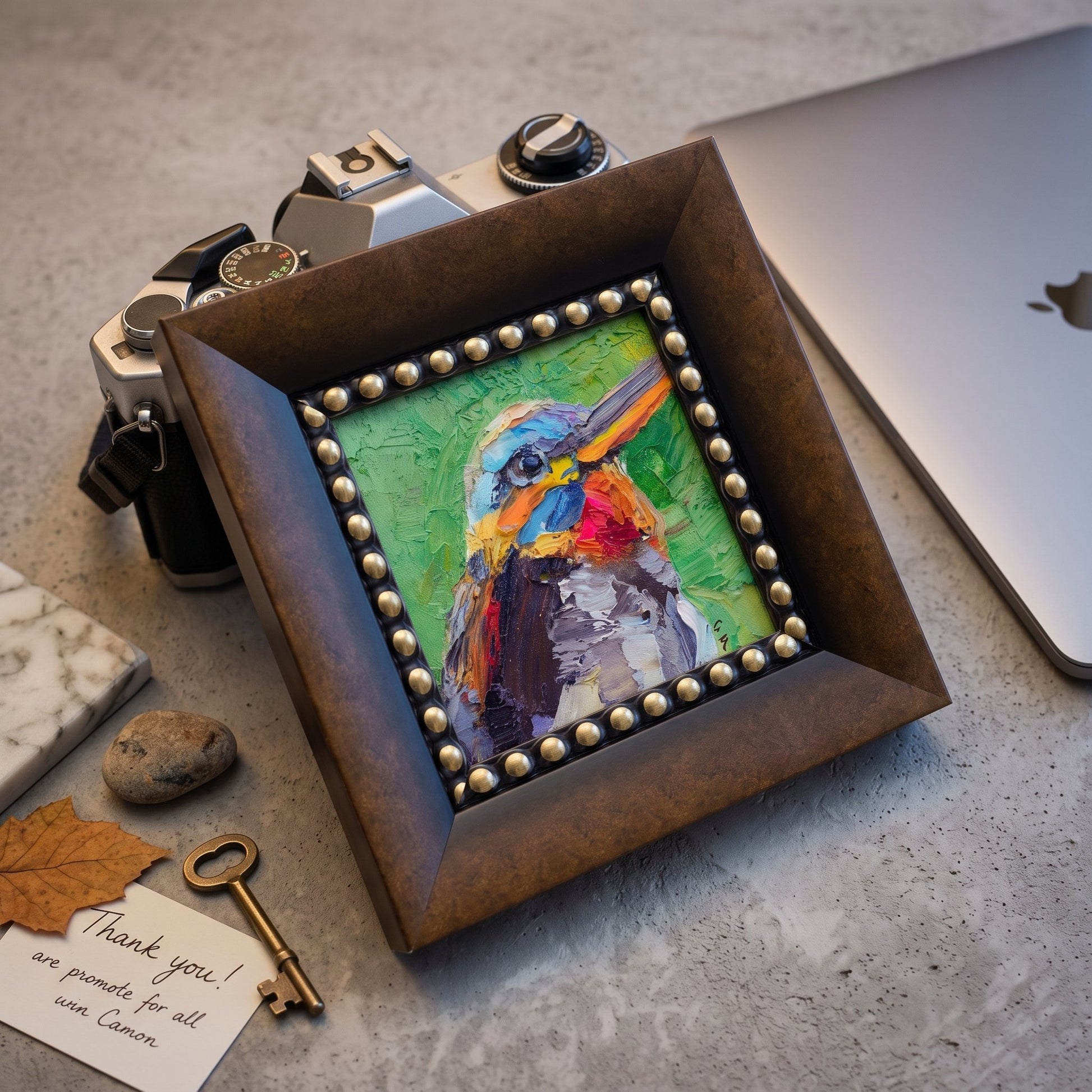 A colorful expressionist bird painting on canvas, framed in ornate metal with beaded trim, sits beside a vintage camera and laptop on a concrete desk.