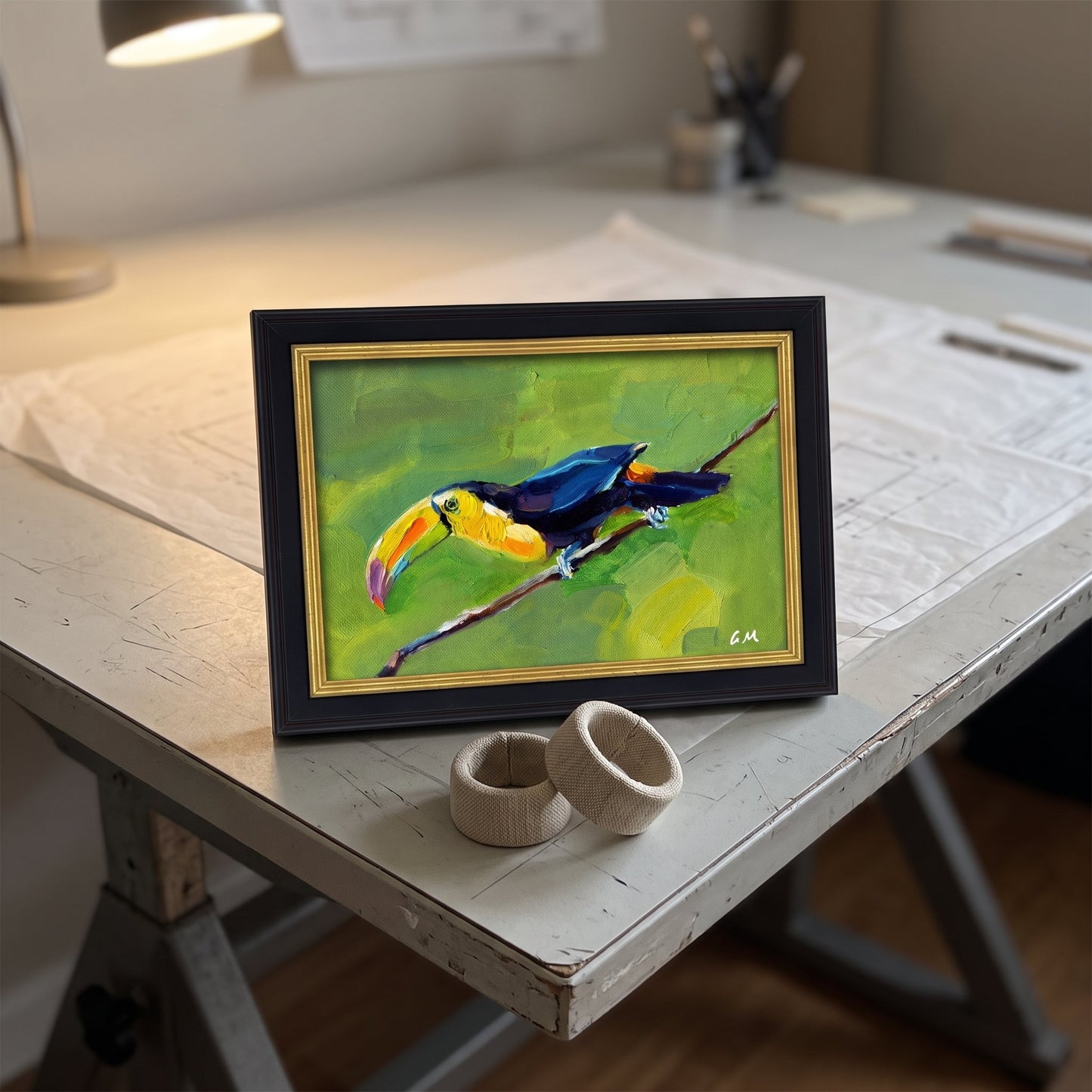 A colorful toucan painting in a black and gold frame rests on a rustic worktable, illuminated by warm desk light.