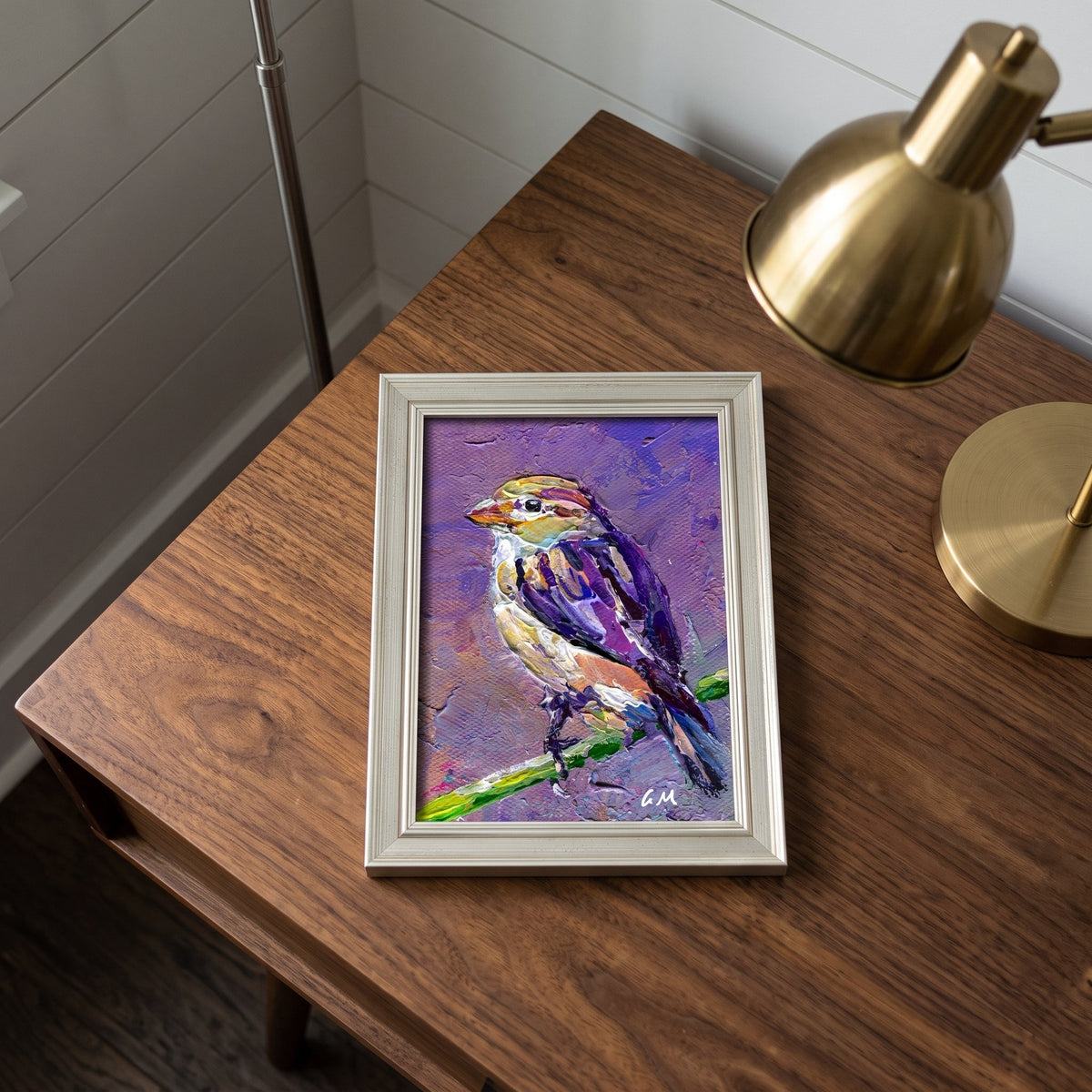 A colorful bird painting in purple, yellow, and white rests on a wooden desk beside a brass lamp, framed in silver.