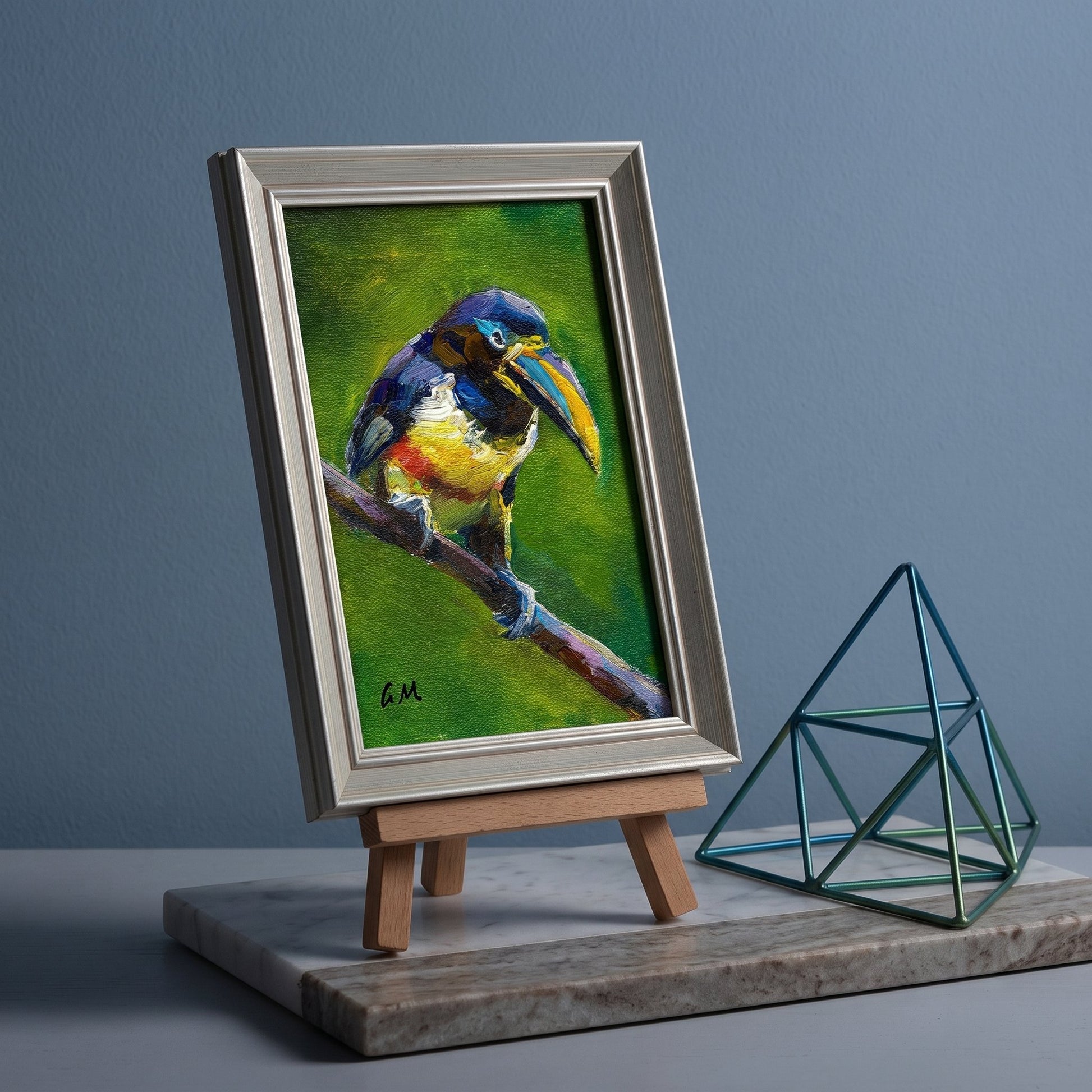 A colorful toucan painting in a silver frame sits on a wooden easel beside a teal geometric sculpture on a marble tray.