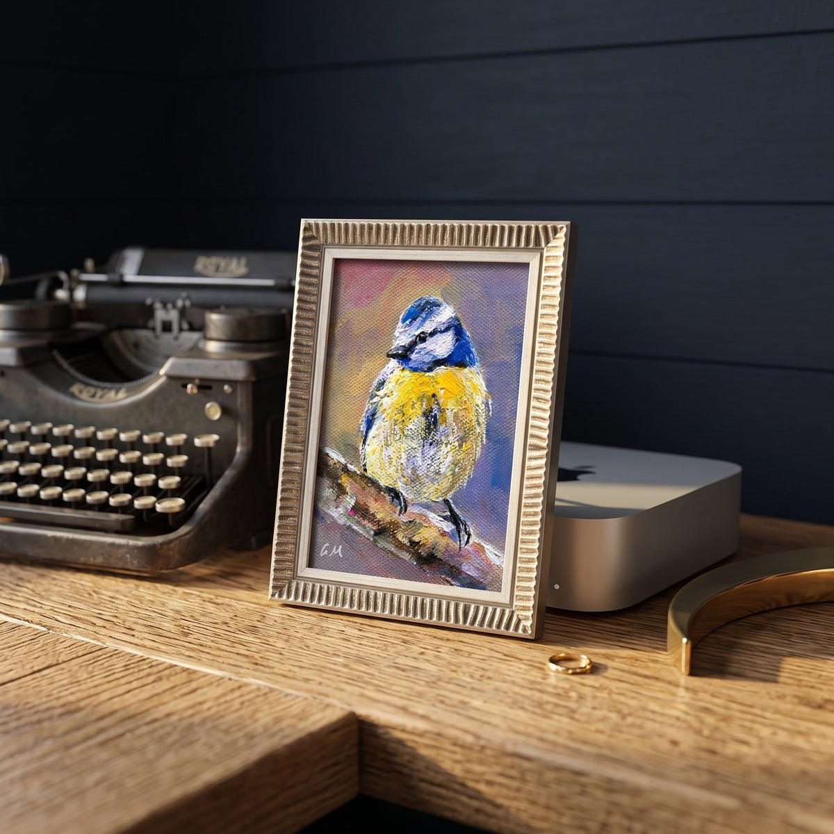 A vibrant blue and yellow bird painting rests on a wooden desk beside a vintage typewriter, framed in gold with textured detailing.