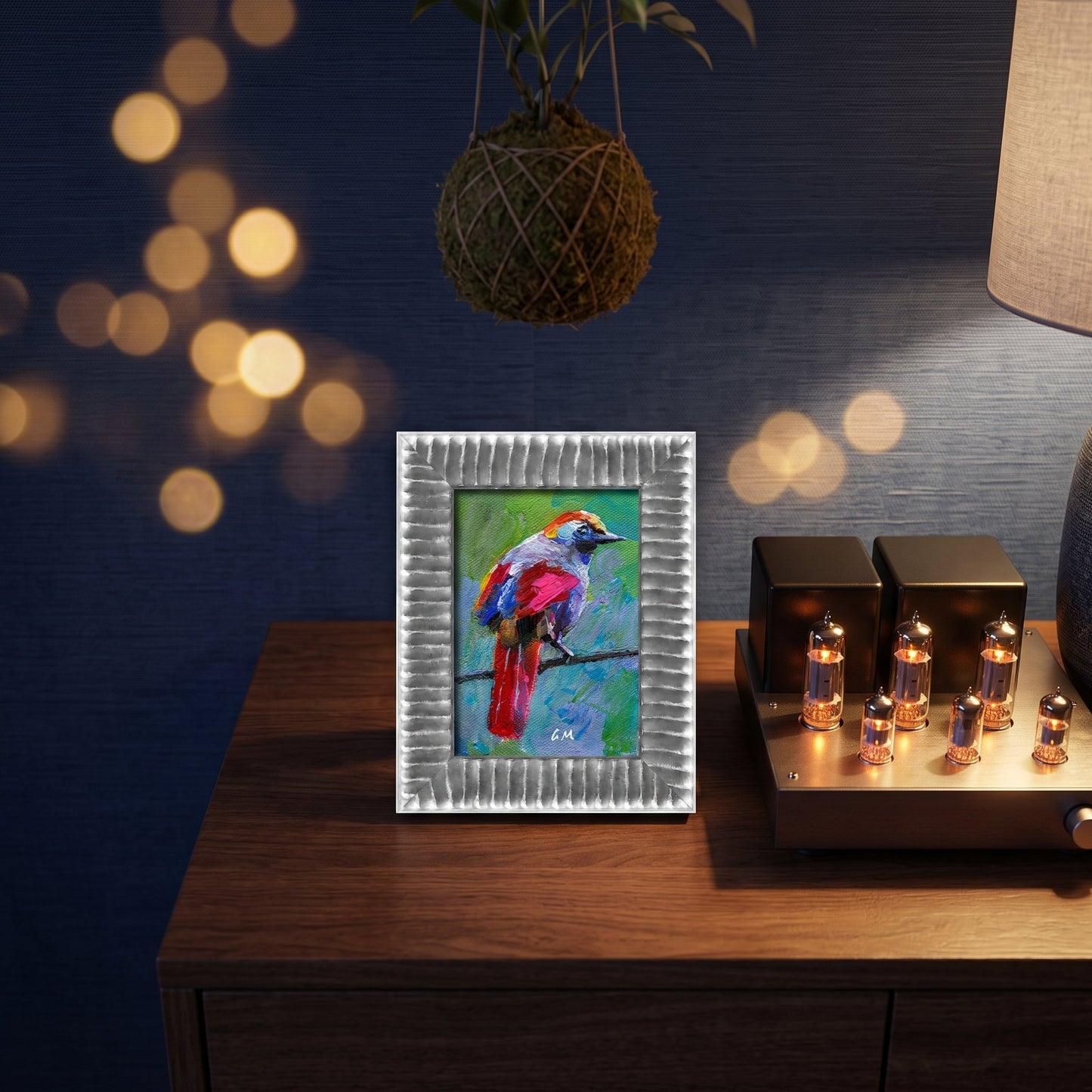 A colorful expressionist bird painting perched on a wooden console beside glowing bulbs and a hanging plant, framed in silver.